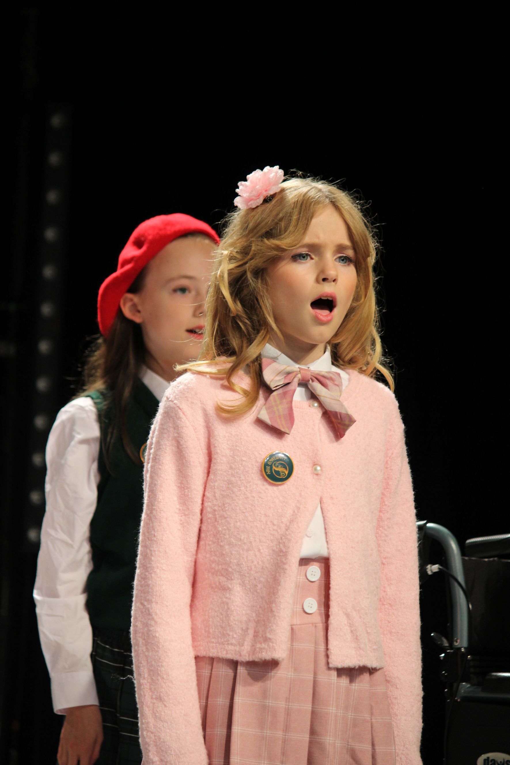 Two young girls singing on a stage. One in pink sweater, the other in a green vest and red hat.