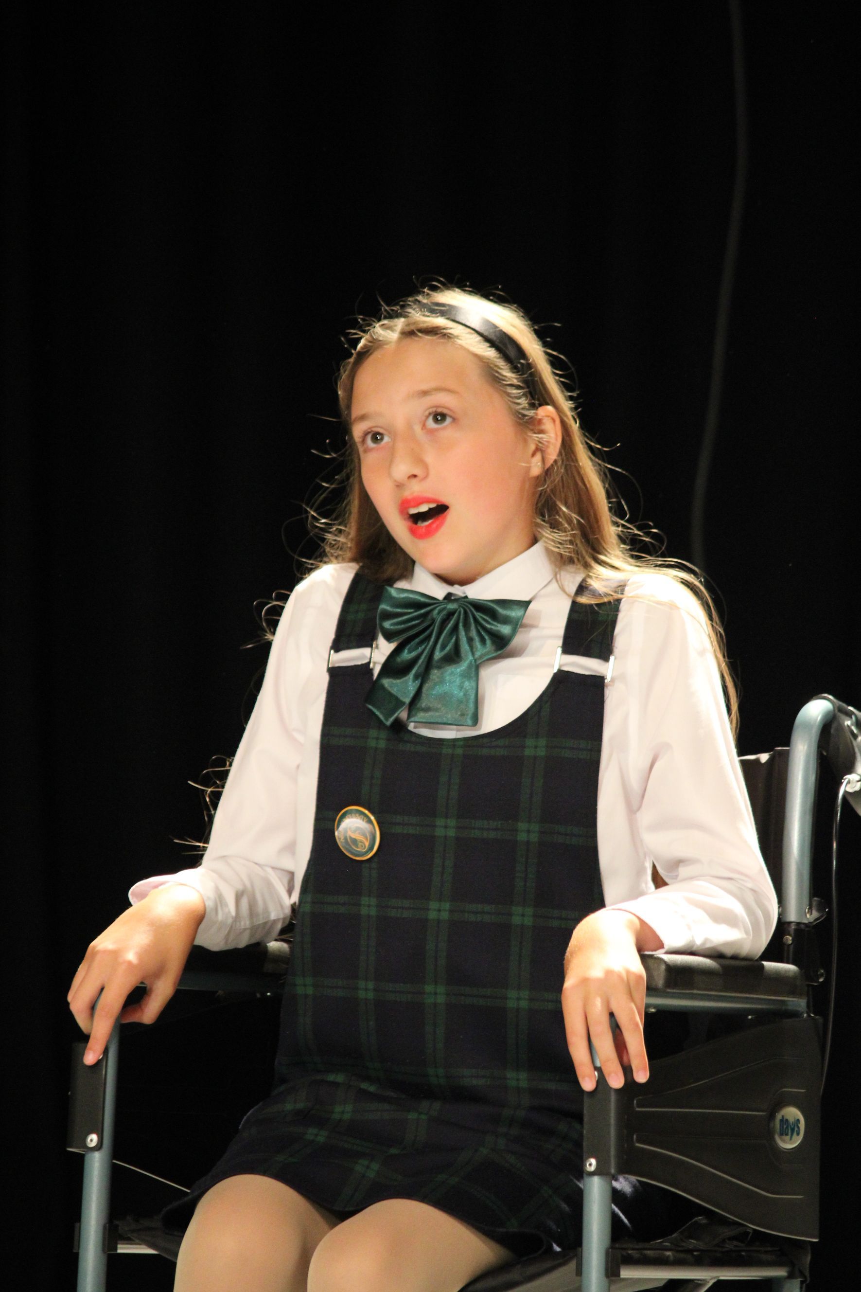 Young person in plaid dress and wheelchair singing, black background.
