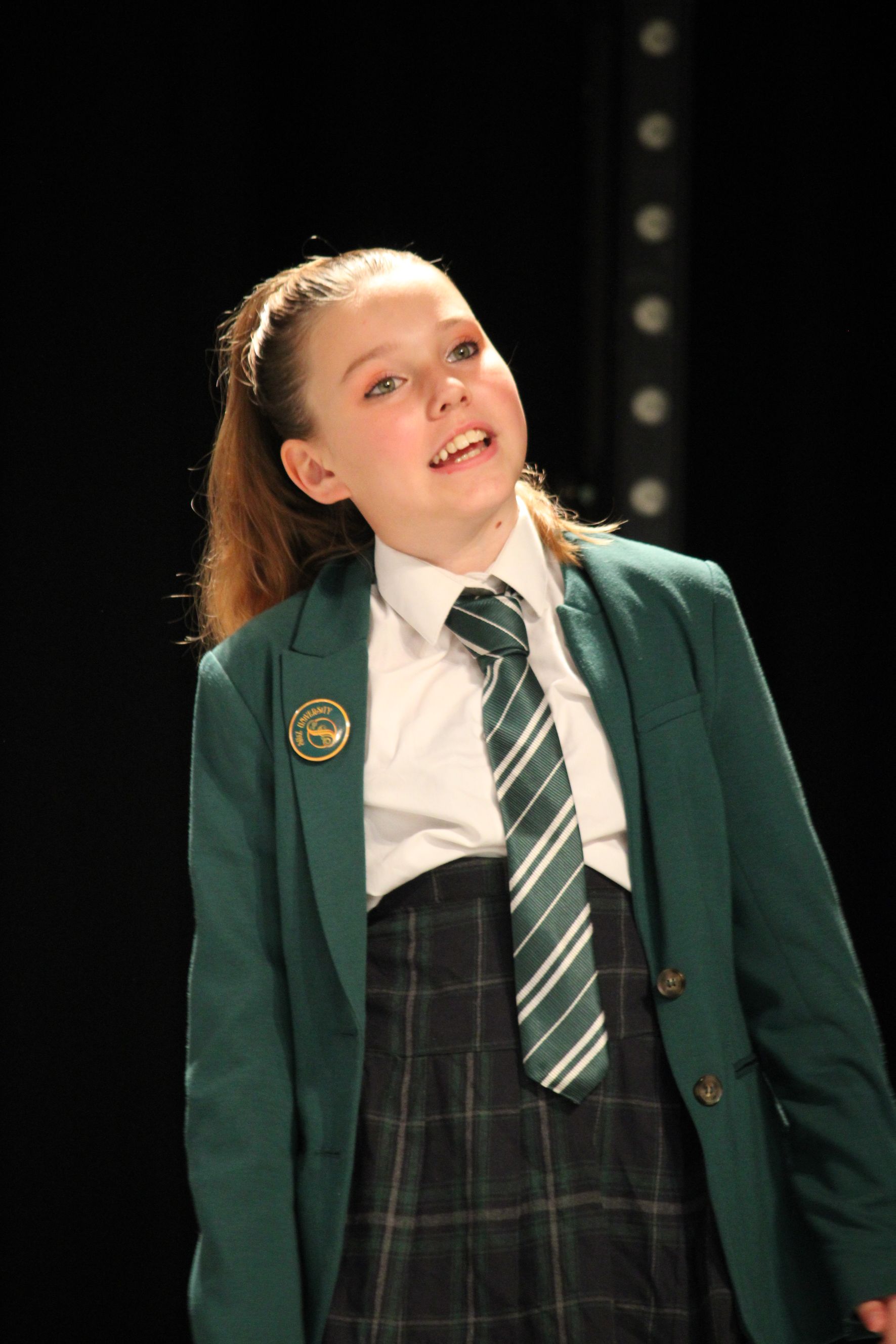 Young person in school uniform singing on stage. Green blazer, tie, plaid skirt, and ponytail. Black background.