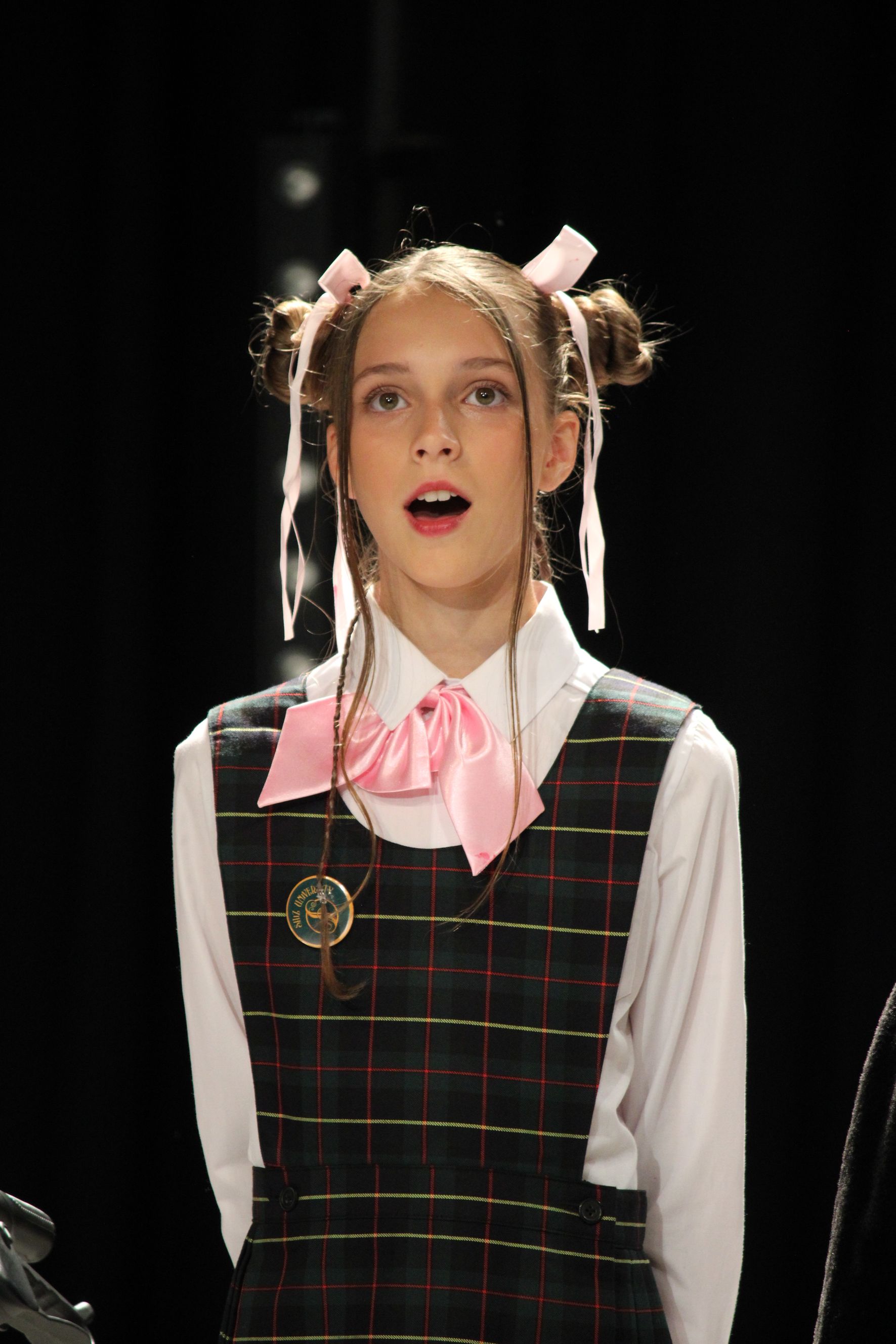 Girl with hair buns, ribbons, school uniform, singing, black background.