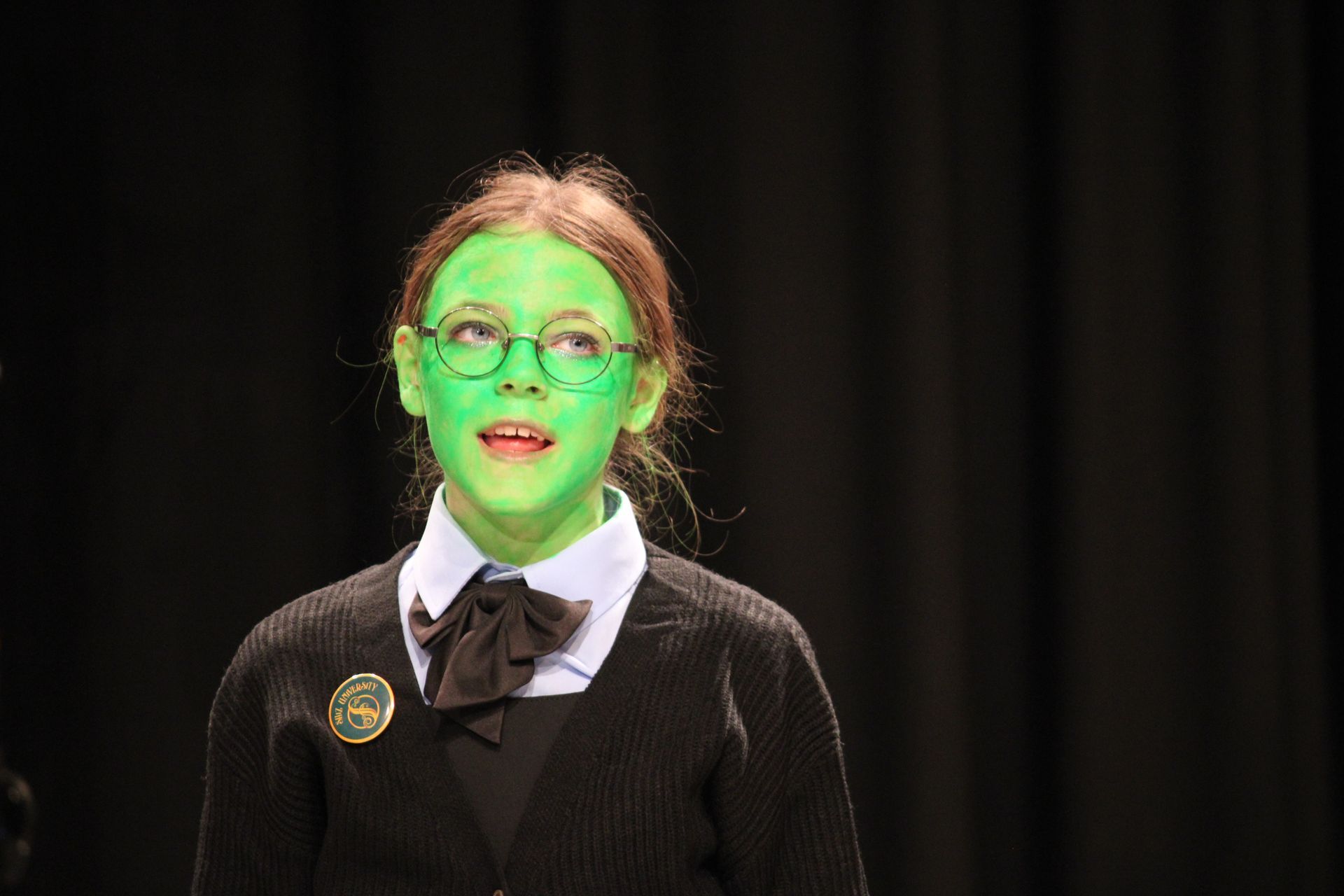 Child with green face paint, glasses, and school uniform, on a stage with a black backdrop, looking upwards.