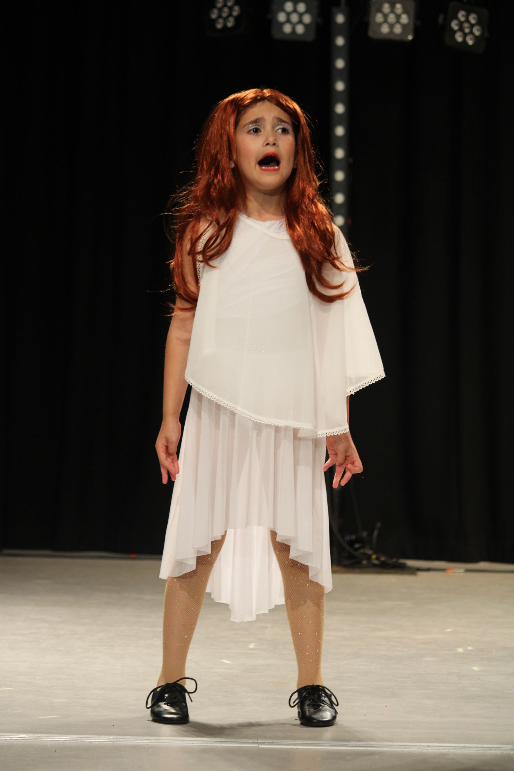 Young performer in a white dress and red wig sings on stage with mouth open. Black background.