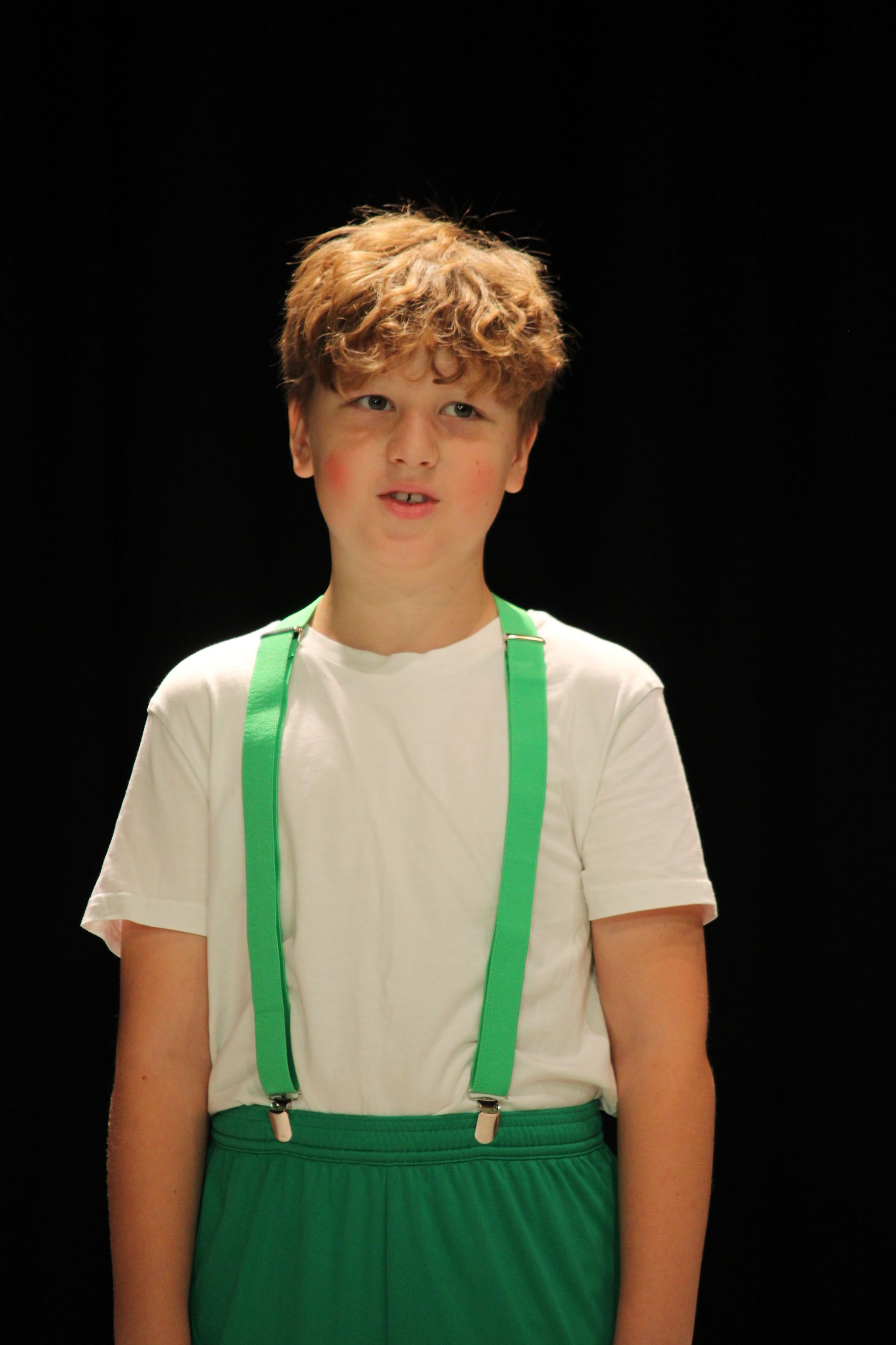 Boy with curly hair, in white shirt, green pants and suspenders, on black backdrop.