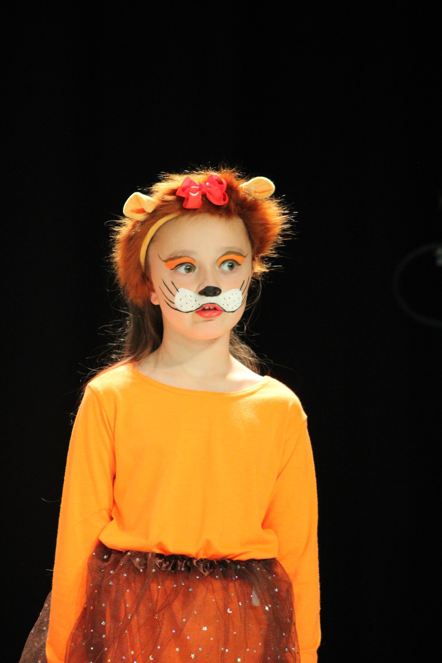 Child in a lion costume on stage, wearing orange top, brown tutu, and face paint.