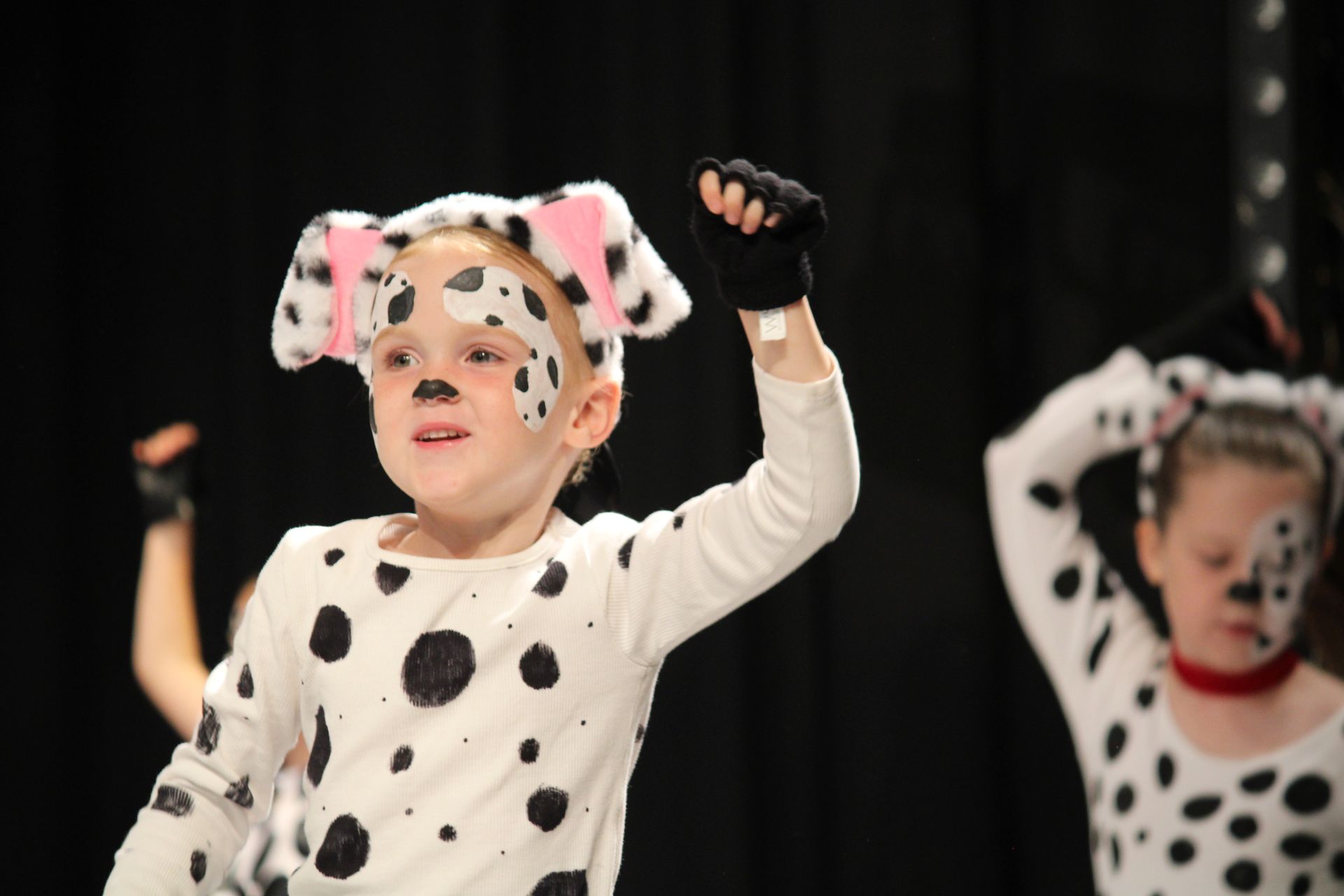 Child in Dalmatian costume, arms raised, performing on stage. White spotted outfit, black gloves, ears, face paint.