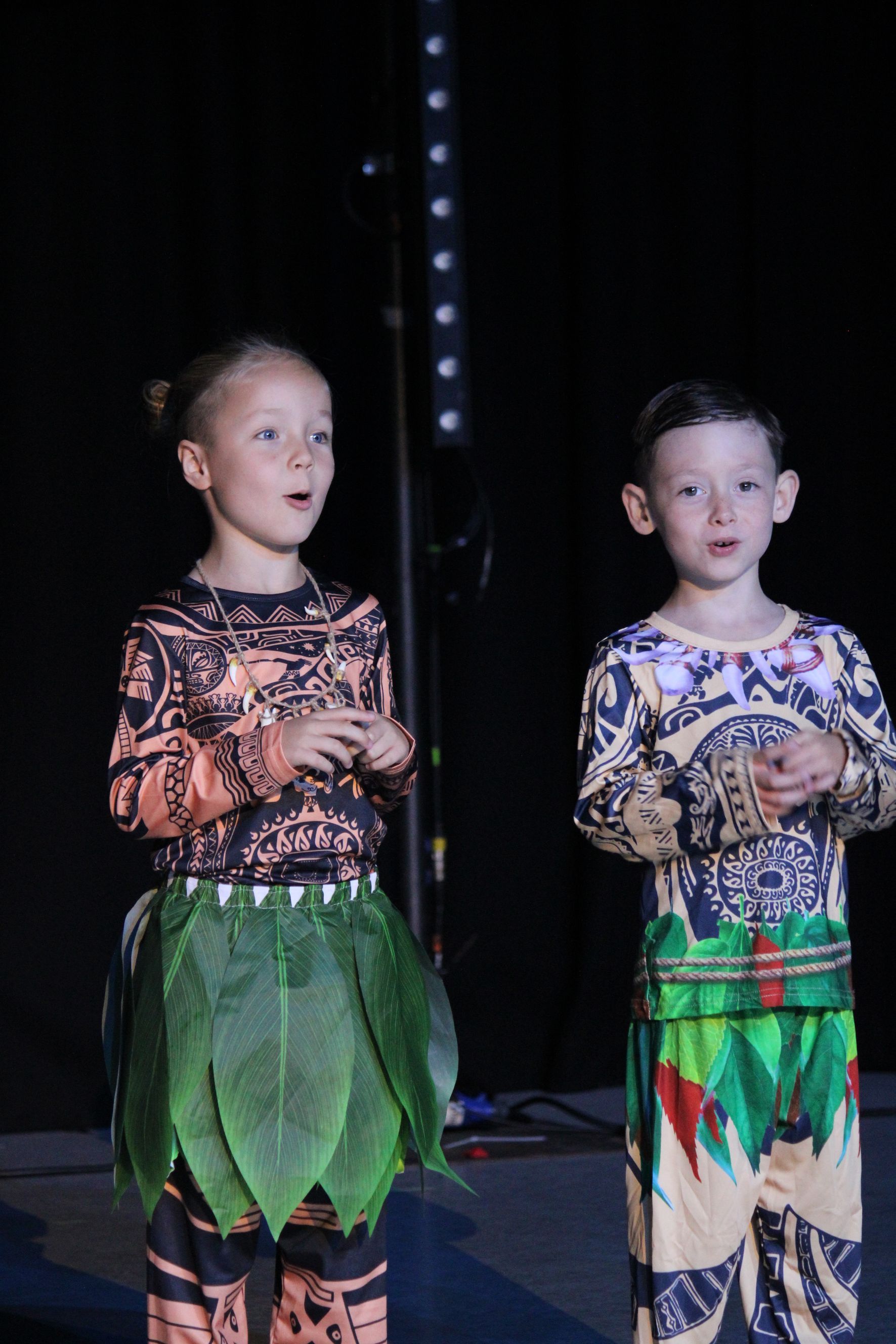 Two children in colorful costumes performing on stage; one has leaf skirt.