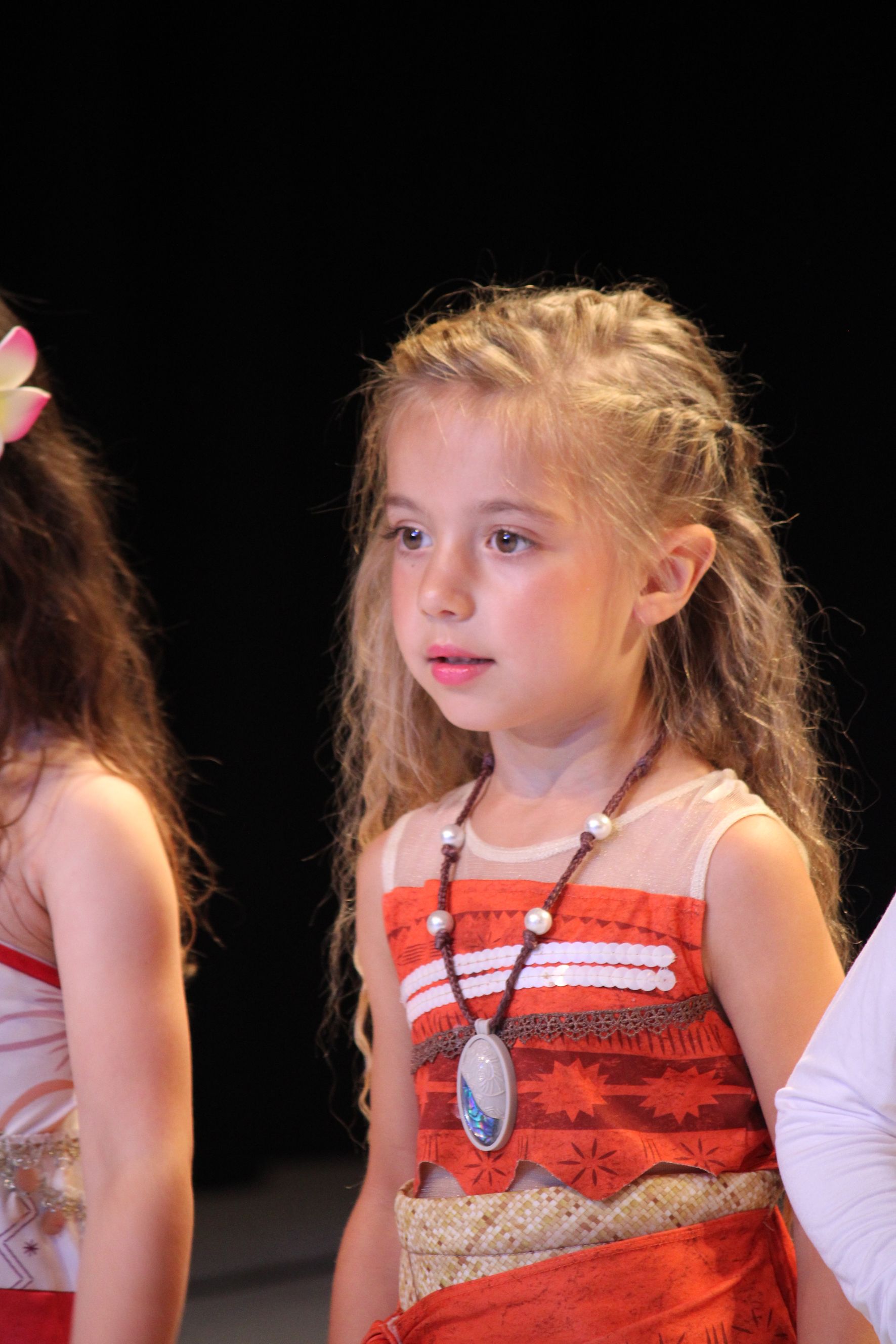 Girl in a Moana costume with necklace, long wavy hair, and serious expression.