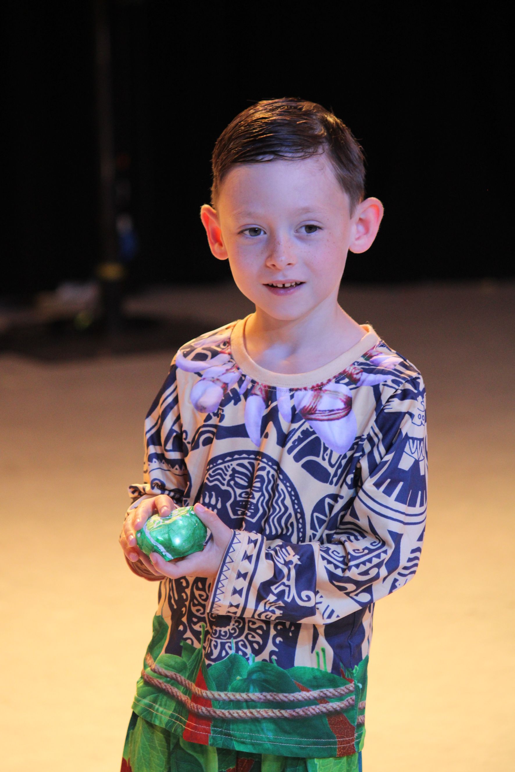 Young child smiling, holding green objects, wearing patterned top and green pants, on stage.