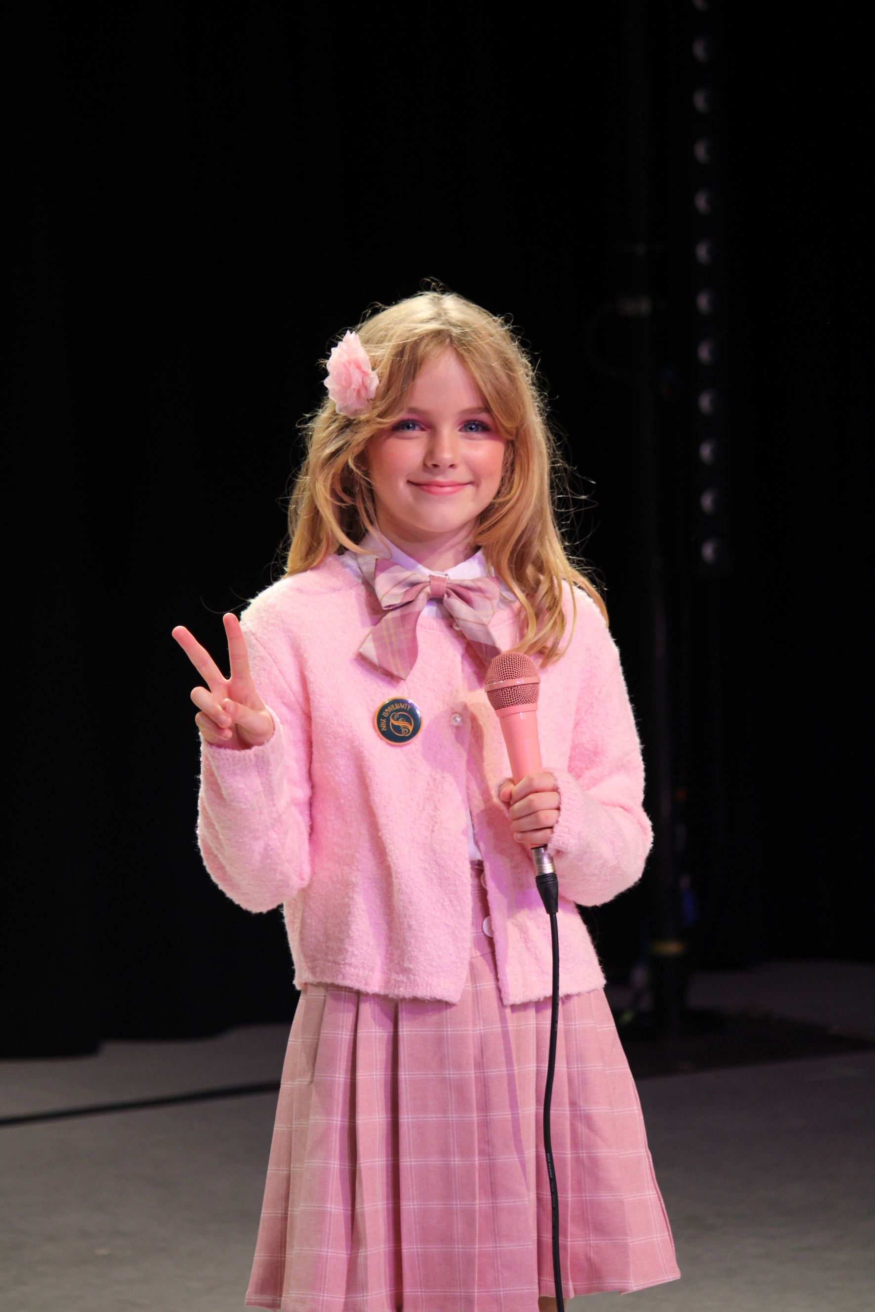 Girl in pink outfit holding a microphone, making a peace sign, and smiling against a dark background.