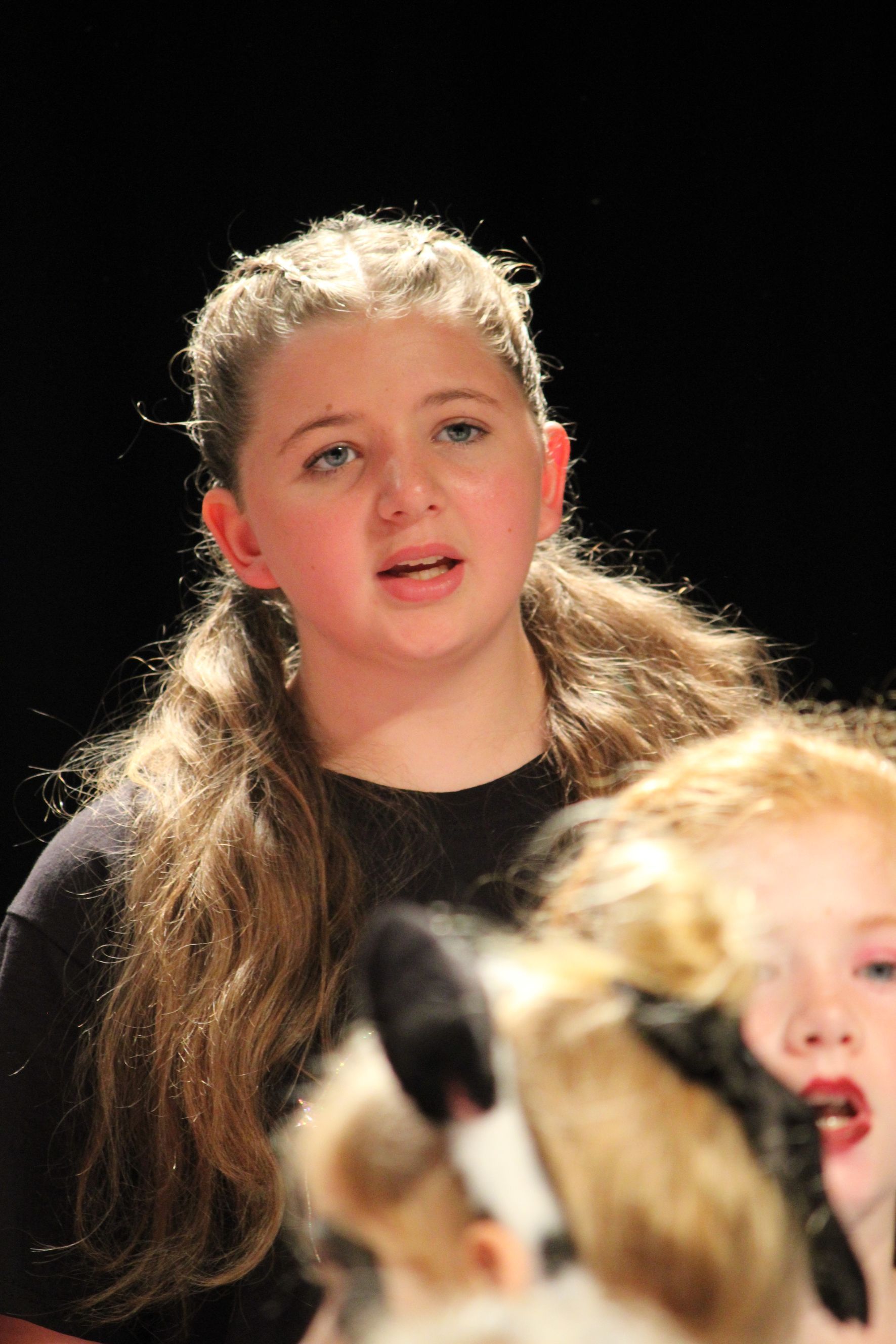 Girl singing onstage with long hair, dark top. A blurred figure with a mask is in front.