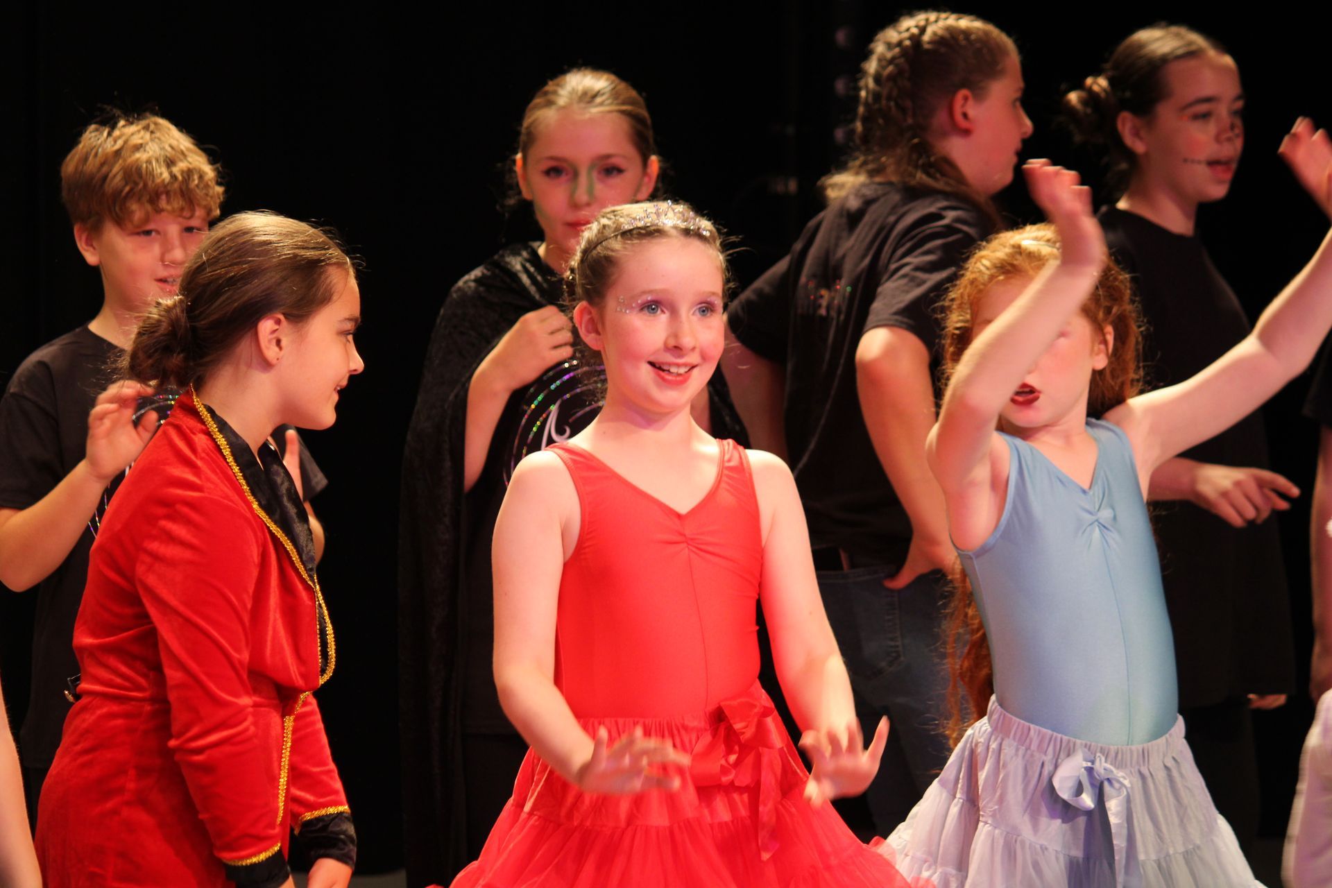 Children in a stage production, some in costume, performing on stage with happy expressions.