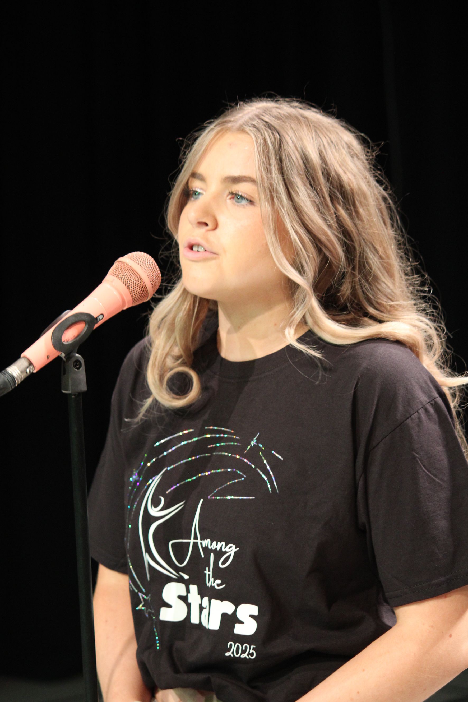Woman with blonde hair speaks into a microphone on a stage. She wears a black t-shirt.