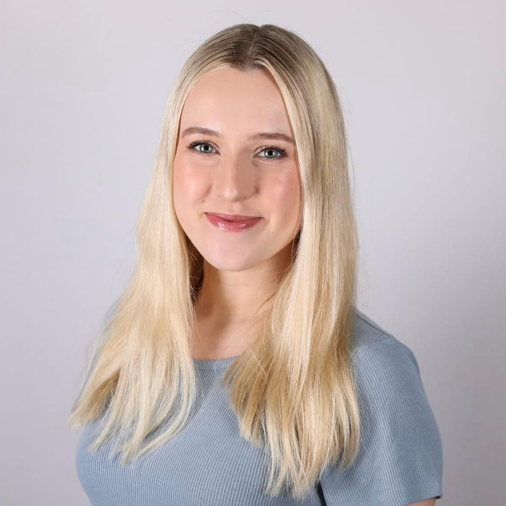 Blonde woman with long hair smiling, wearing a blue top, against a gray background.