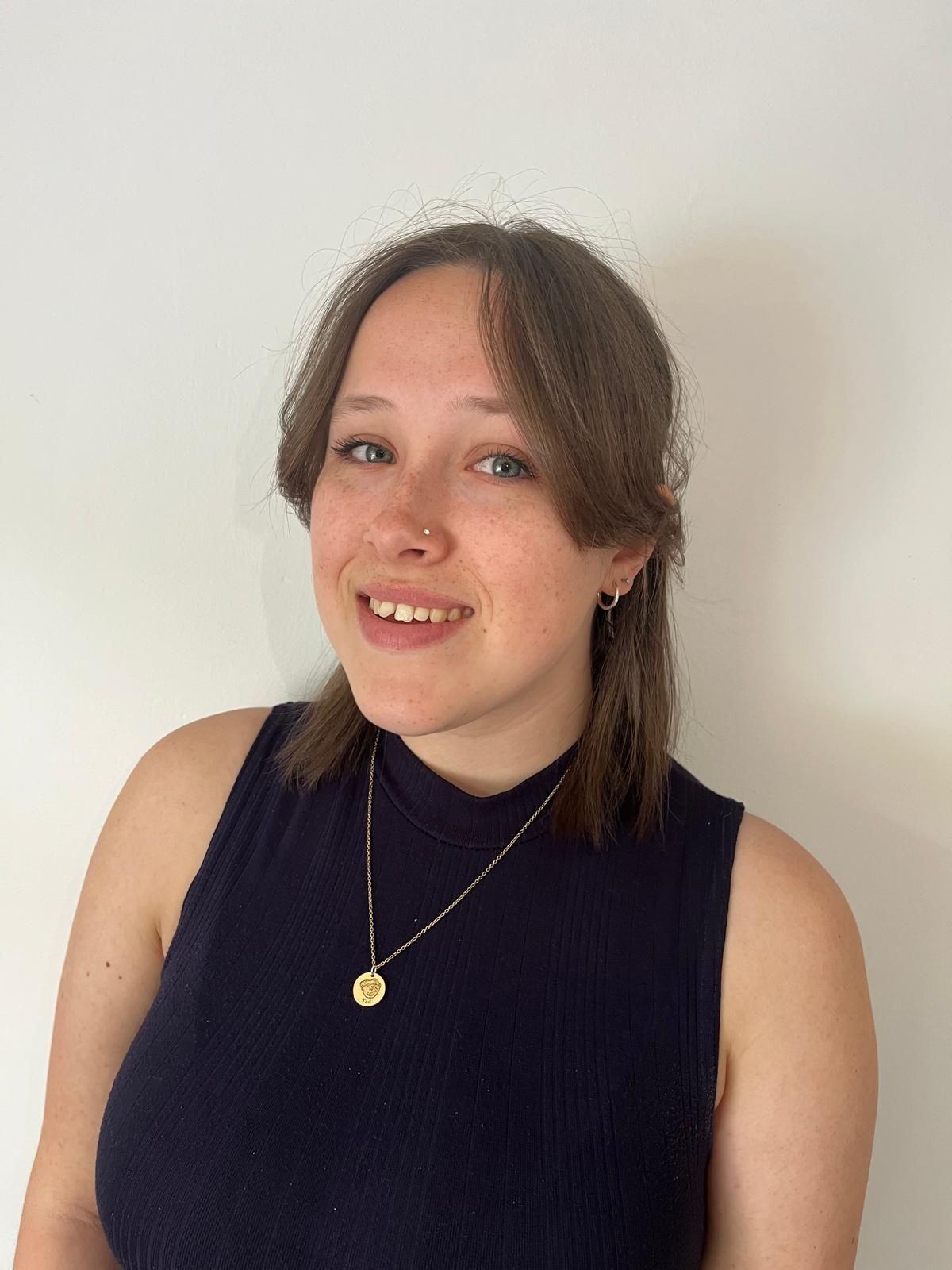 Woman with brown hair smiles, wearing a navy tank top and gold necklace against a white wall.