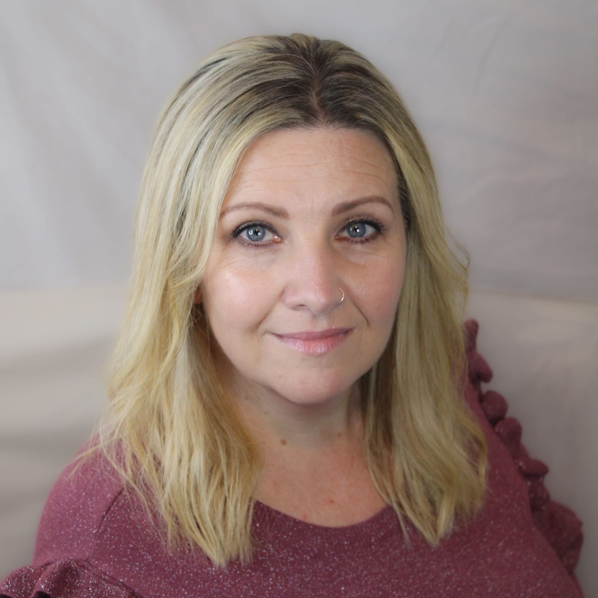 Blond woman with blue eyes smiles, wearing a pink shirt, in front of a white backdrop.