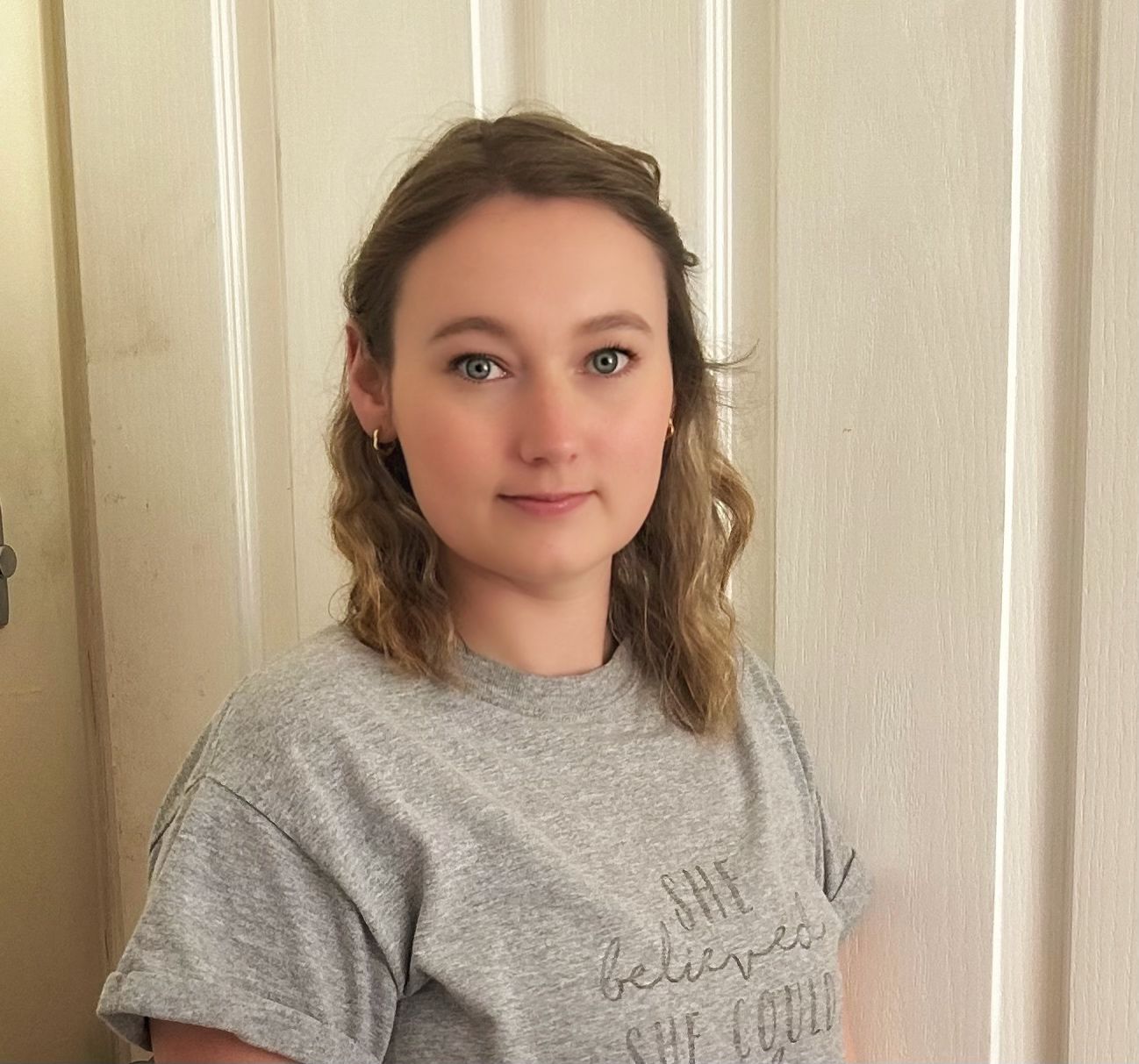 Woman with light brown hair in grey t-shirt stands in front of a white door.