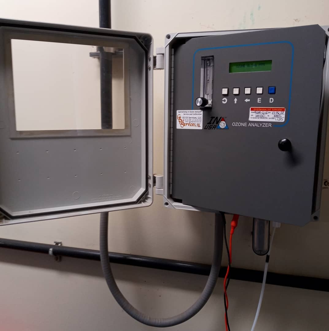 Wall-mounted industrial controller box with a digital display, buttons, and a flow meter, open to reveal interior controls.