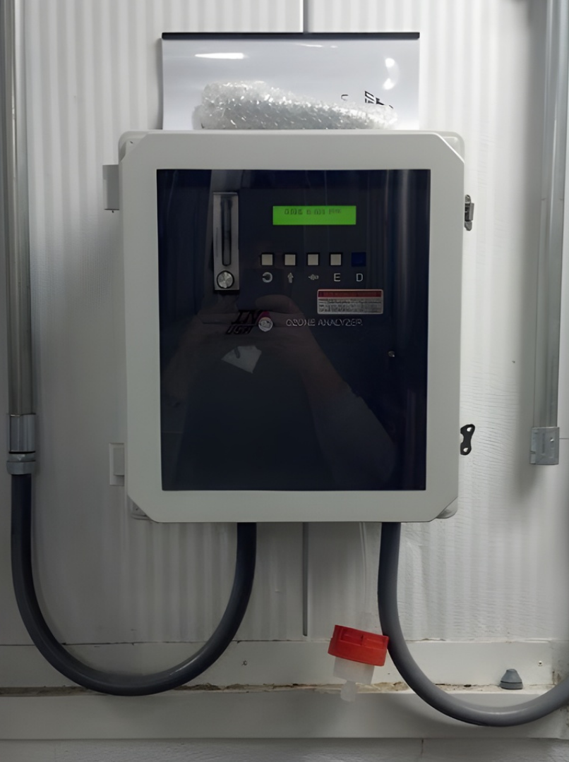 A close-up of a gray, wall-mounted industrial digital monitor with white tubing connections in a factory setting.