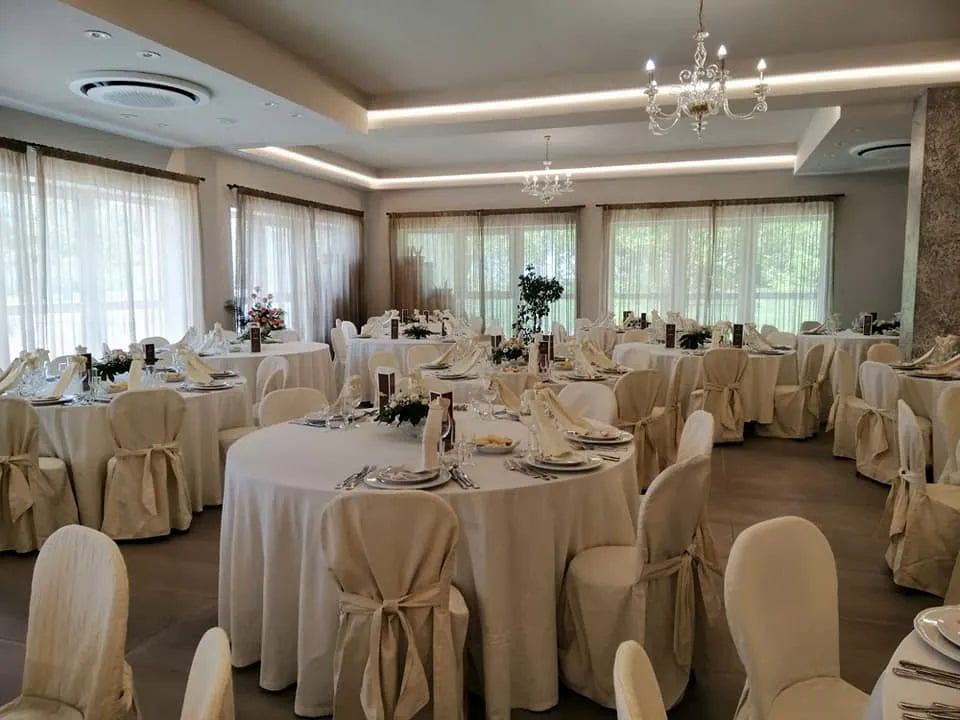 Elegante sala per banchetti allestita per un matrimonio. Tavoli rotondi con tovaglie e coprisedie bianchi, luci soffuse.