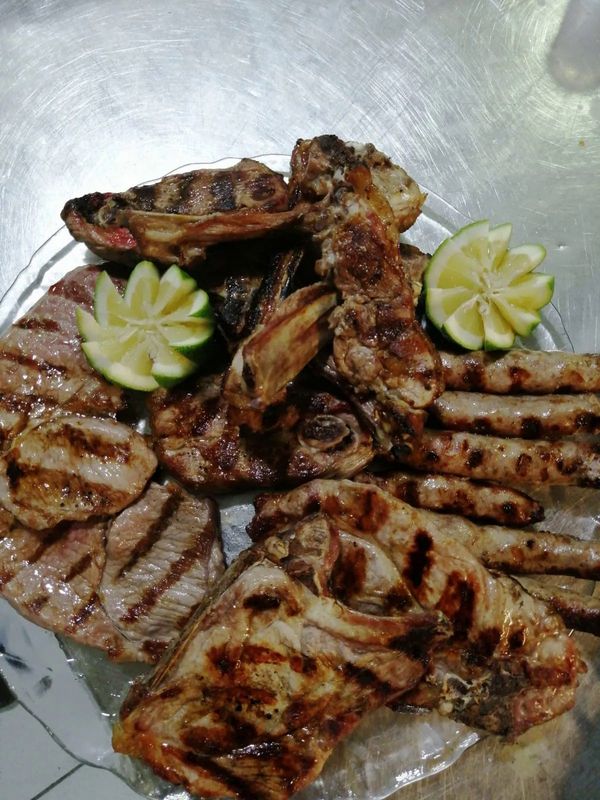 Carni grigliate, tra cui costolette e salsicce, servite su un piatto, guarnite con spicchi di lime.