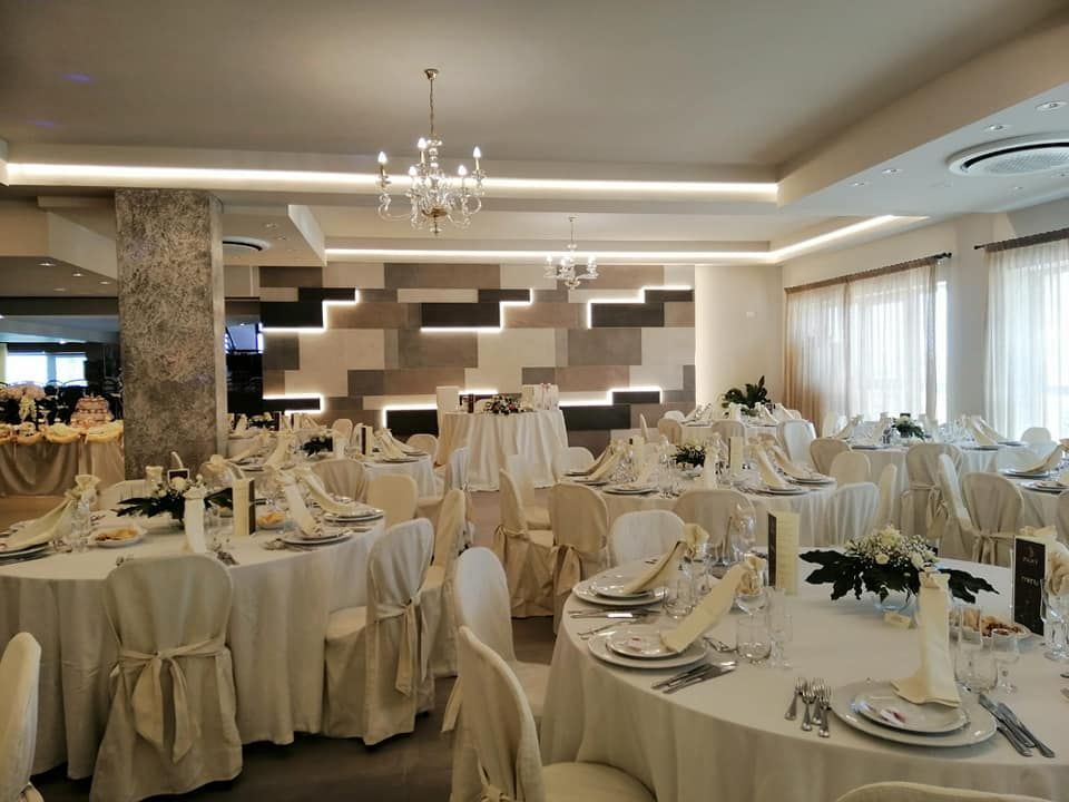 Sala da pranzo formale allestita per un matrimonio, con tavoli rotondi, tovaglie bianche e parete decorativa.