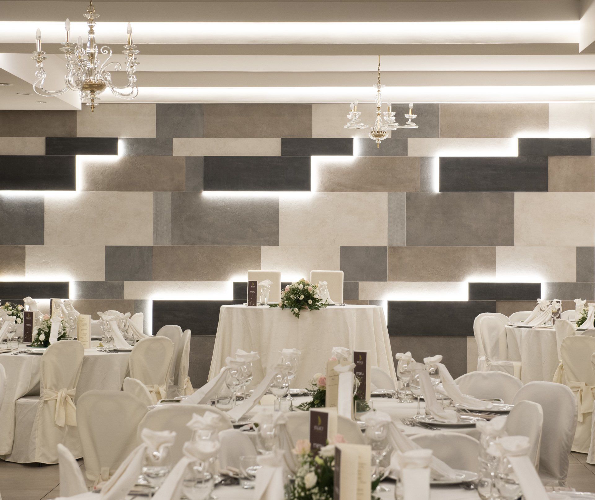 Sala per ricevimenti nuziali, tavoli apparecchiati con tovaglie bianche, sedie e centrotavola, contro una parete decorata con luci da incasso.