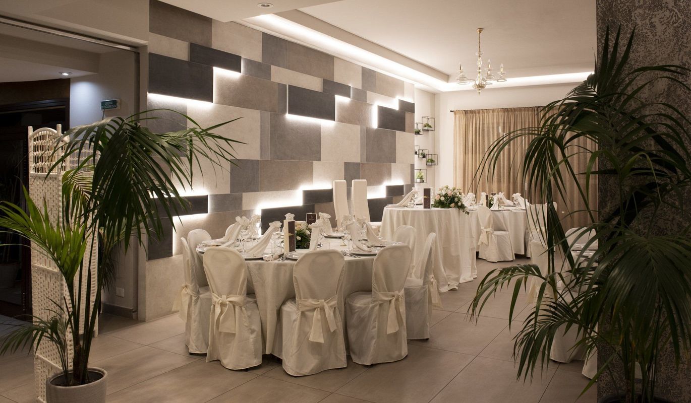 Elegante sala per banchetti con tavoli e sedie bianchi e una parete decorativa. Piante verdi incorniciano la sala.