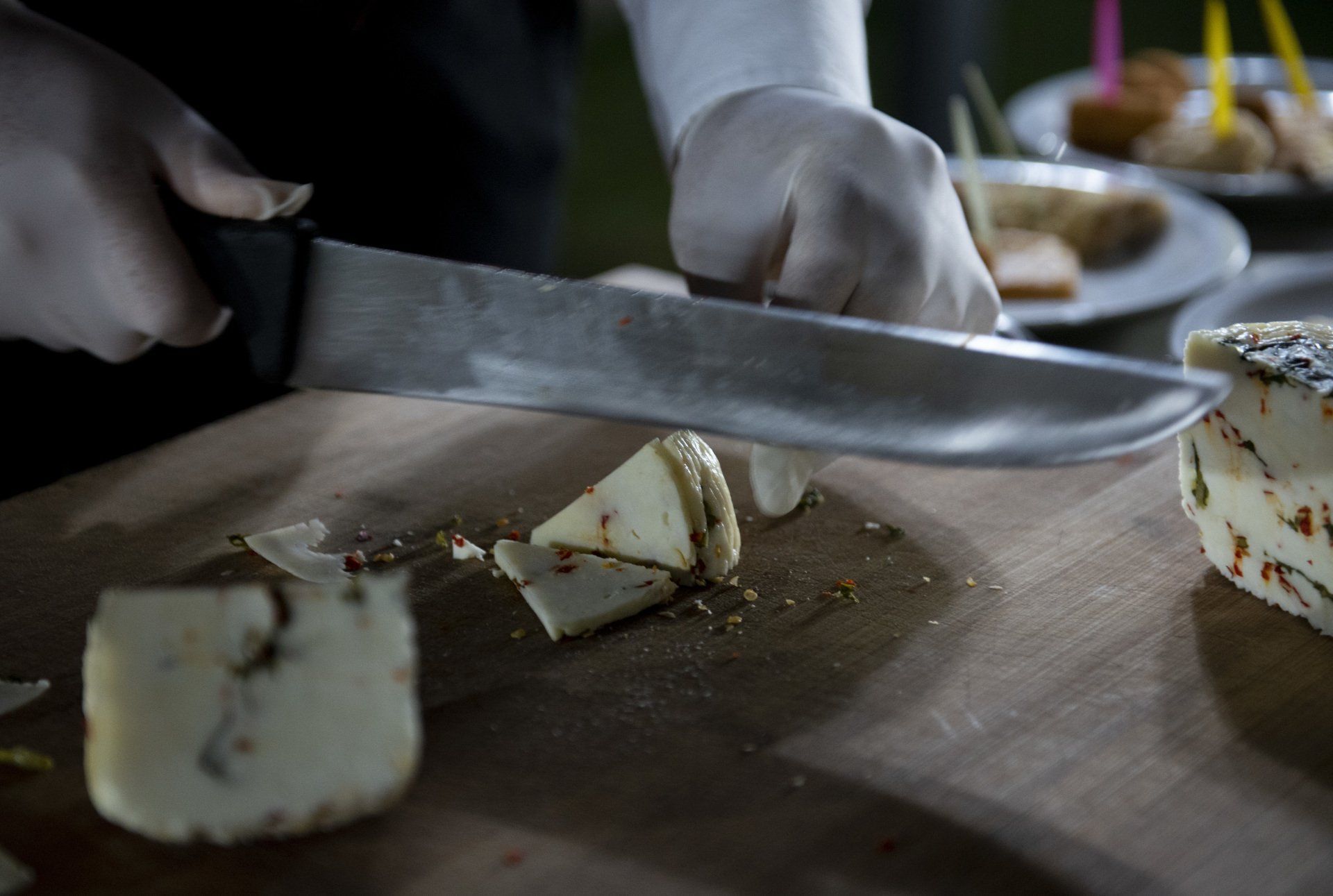 Mani guantate tagliano il formaggio su un tagliere di legno, sullo sfondo altri spuntini.