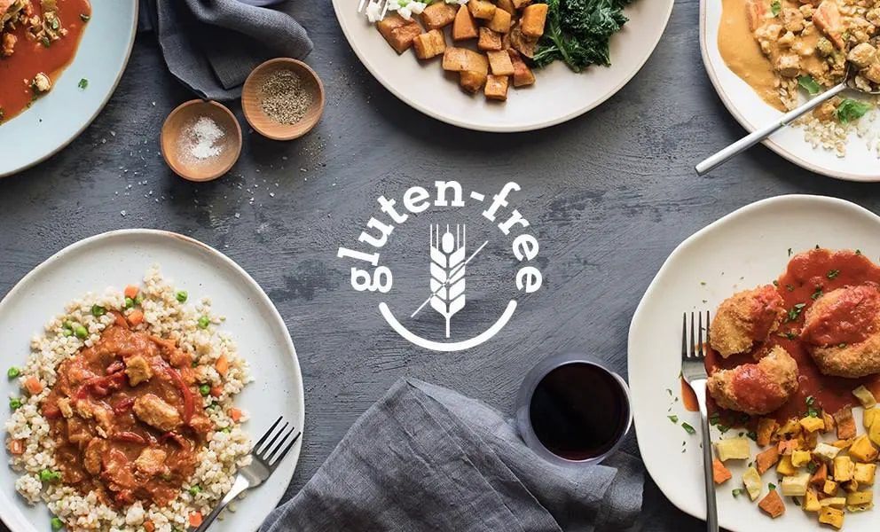 Piatti con pietanze senza glutine, tra cui pollo, verdure e riso, disposti su una superficie grigia.