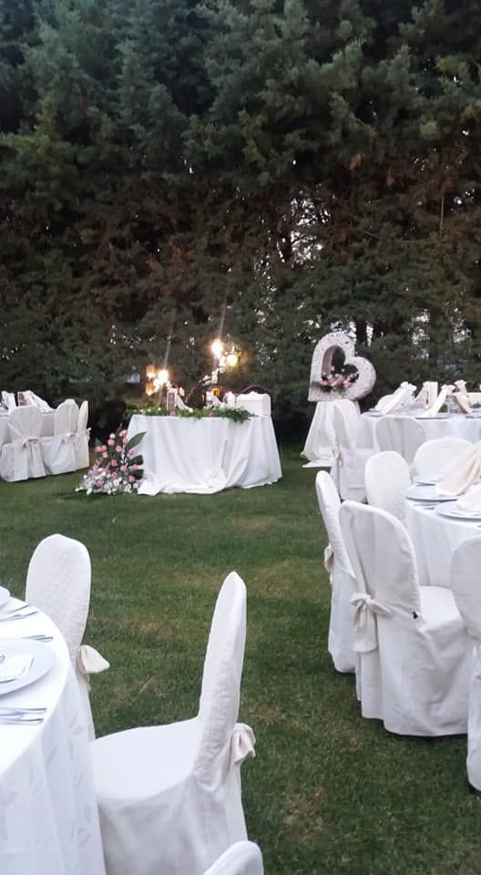 Ricevimento di nozze all'aperto con tavoli, sedie e decorazioni bianche disposte su un prato erboso; sullo sfondo alti alberi verdi.