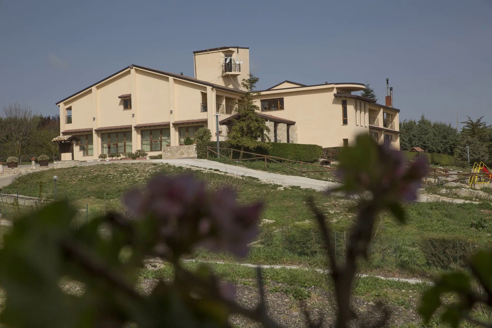 Edificio beige con una torre su una collina erbosa, visto da dietro fiori rosa sfocati in una giornata di sole.