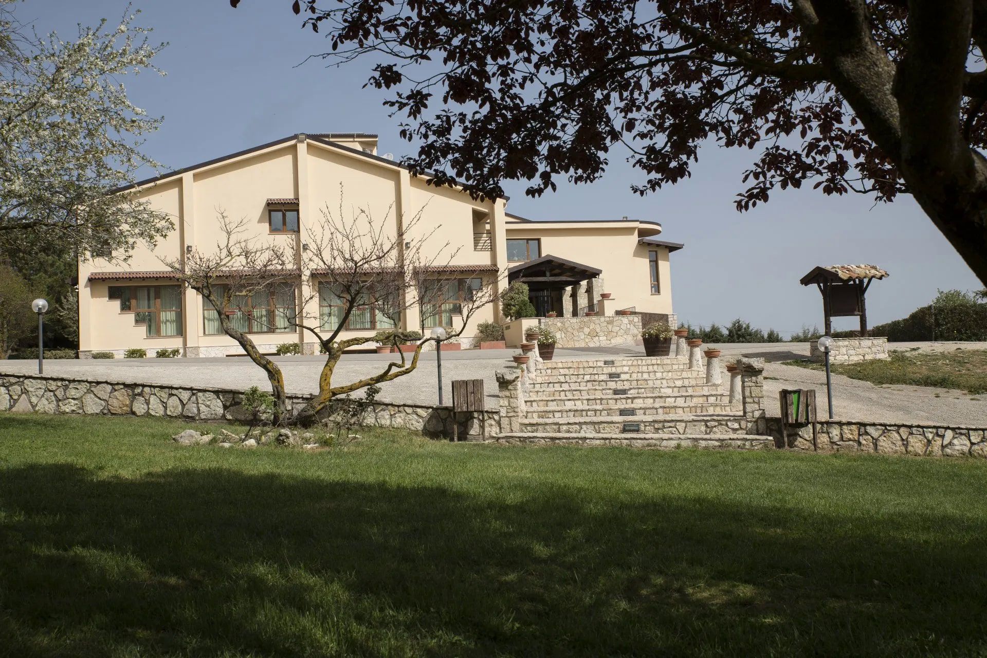 Un edificio giallo con scale in pietra, un cortile erboso e alberi in una giornata di sole.