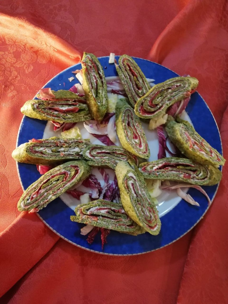 Piadine verdi con ripieno rosso disposte su un piatto con bordo blu, con cavolo rosso su un panno rosso.