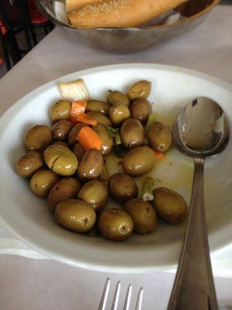 Piatto di olive marroni con verdure e cucchiaio su una tovaglia bianca.