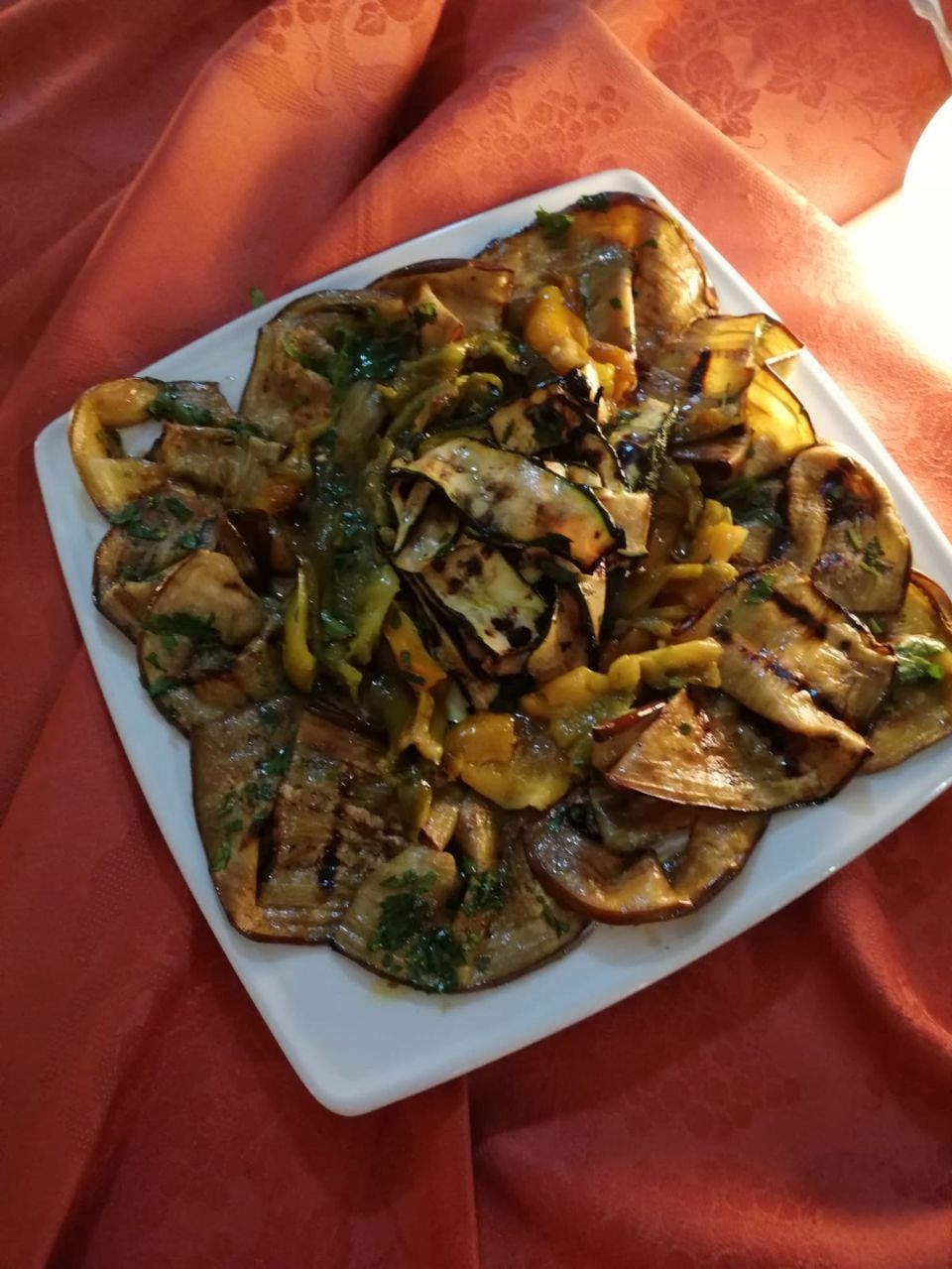 Melanzane e zucchine grigliate su un piatto quadrato bianco, servite su una tovaglia rossa.