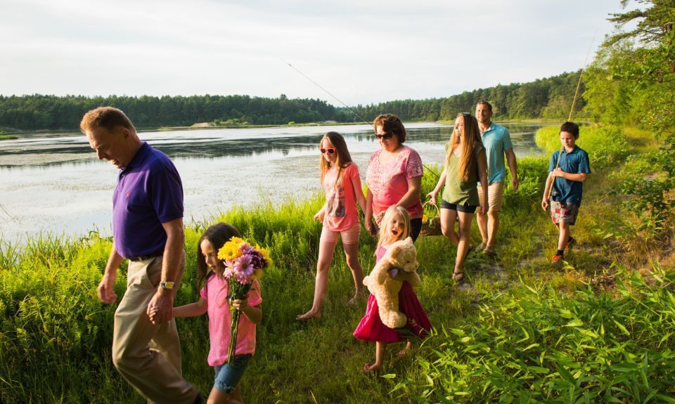 Famiglia che cammina lungo un lago, alcuni con dei fiori in mano. Foglie verdi e acqua.