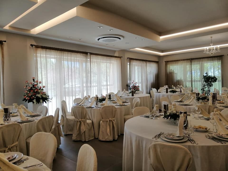 Elegante sala da pranzo apparecchiata per un evento speciale: tavoli rotondi con tovaglie bianche e centrotavola floreali.