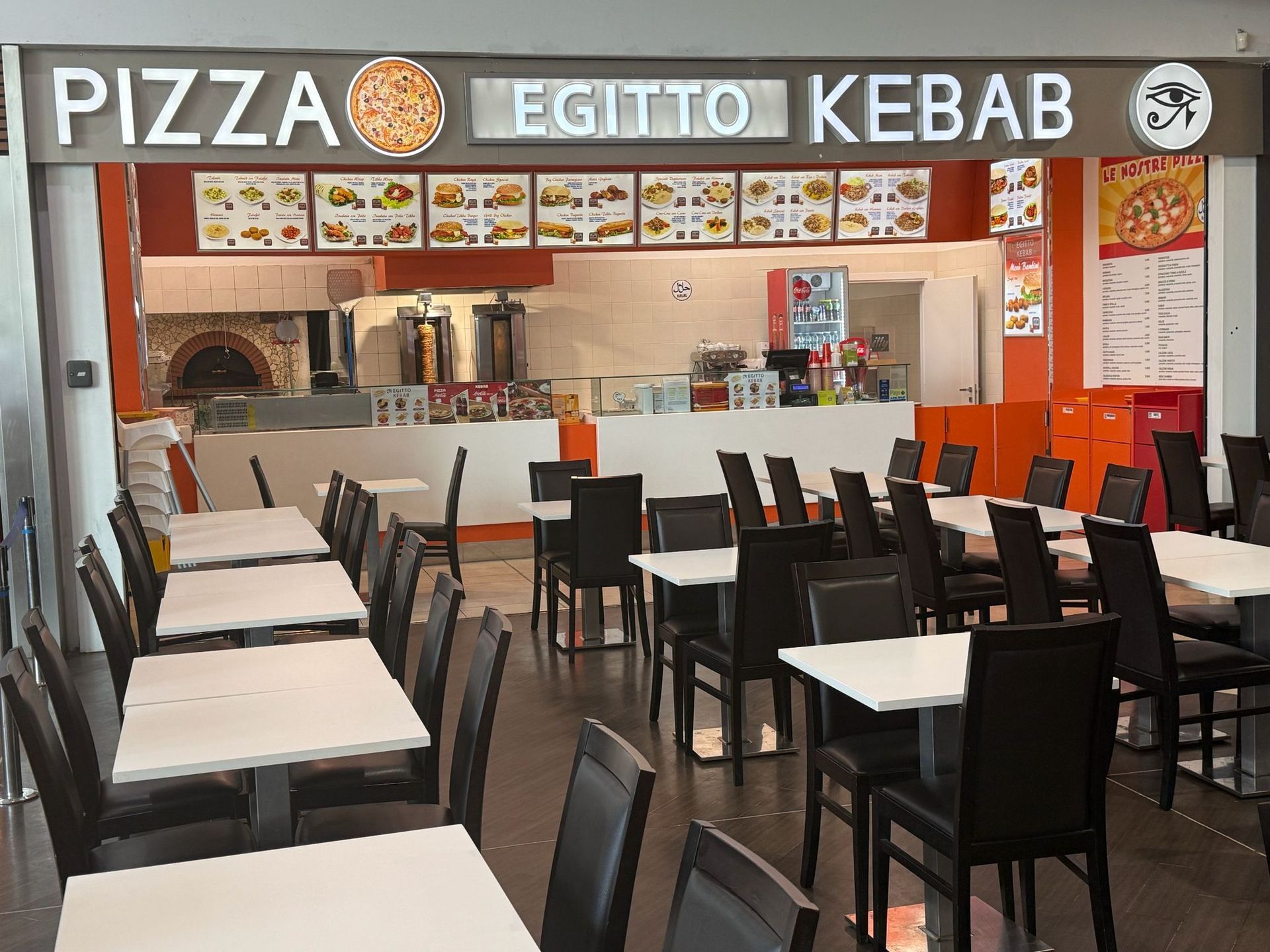 Un ristorante di pizza e kebab vuoto e ben illuminato, con tavoli bianchi, sedie nere e un grande menù esposto sulla parete.
