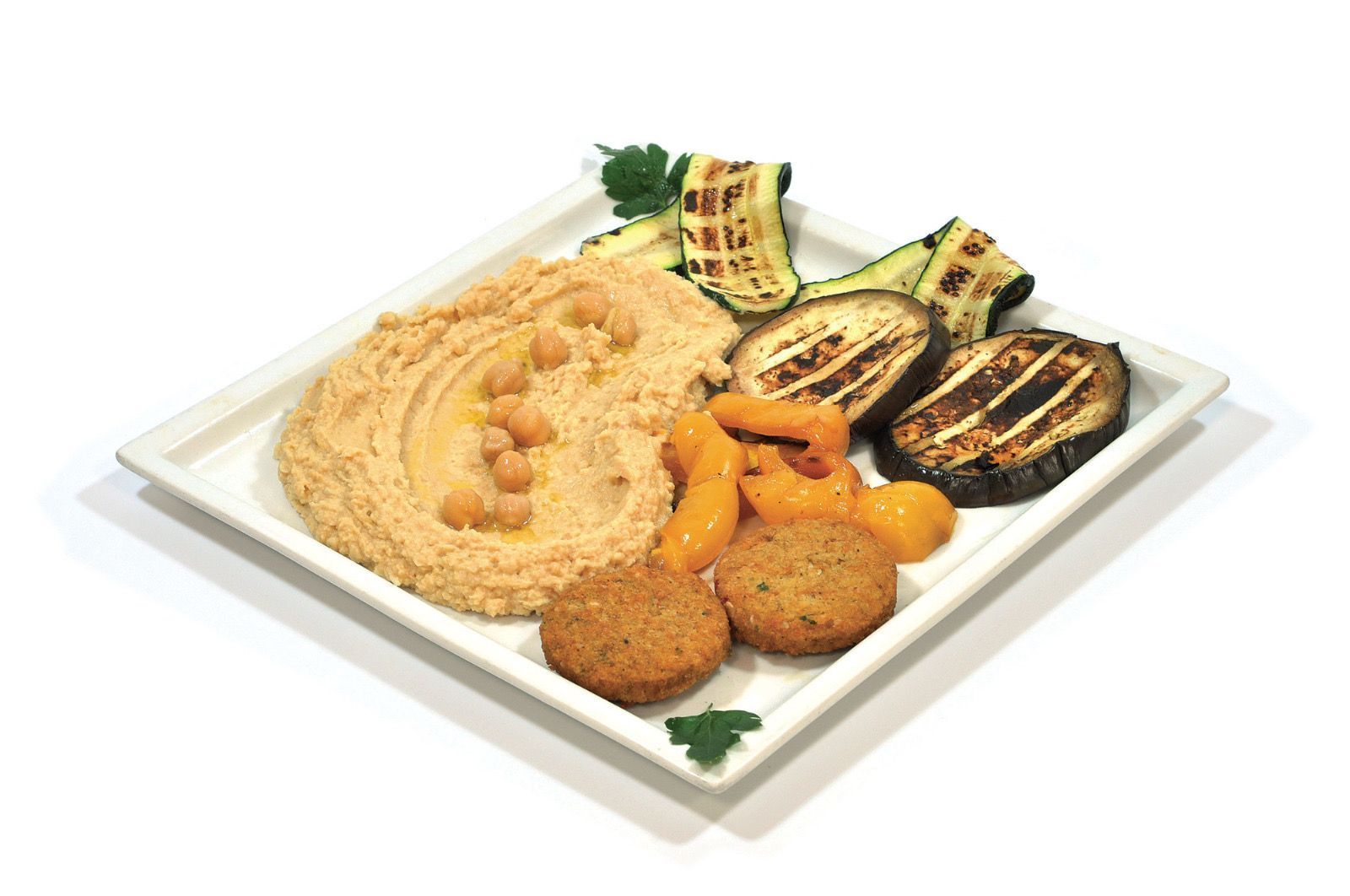 Su un piatto quadrato bianco è disposta una porzione di hummus guarnito con ceci, due polpette di falafel, zucchine grigliate e melanzane.