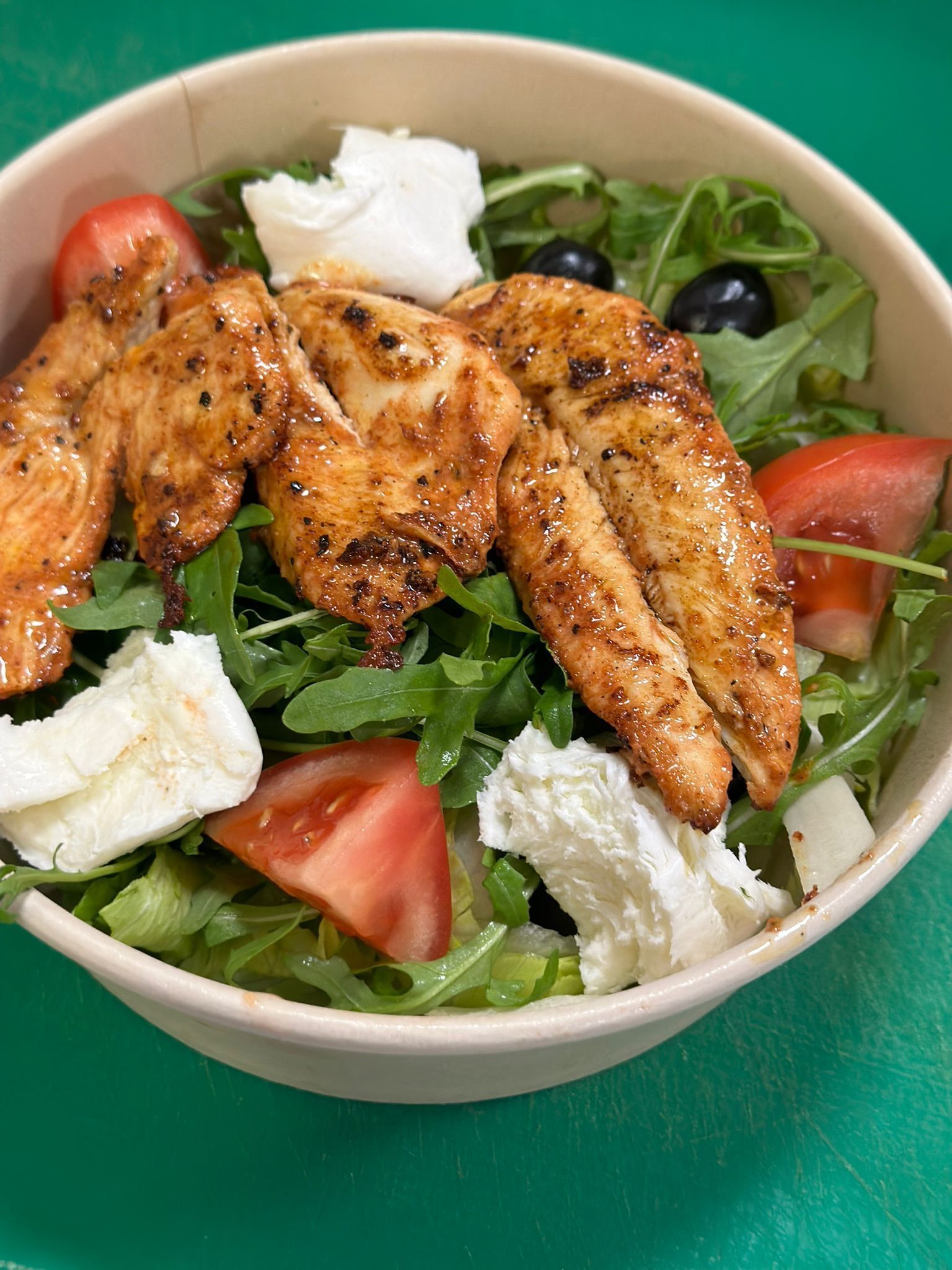 Un'insalata in una ciotola di carta con straccetti di pollo speziati, rucola, spicchi di pomodoro, olive nere e cubetti di formaggio bianco.