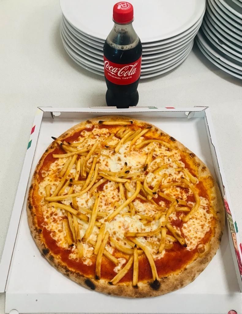 Una pizza condita con patatine fritte in una scatola di cartone bianca, accanto a una bottiglia di Coca-Cola e a pile di piatti bianchi.