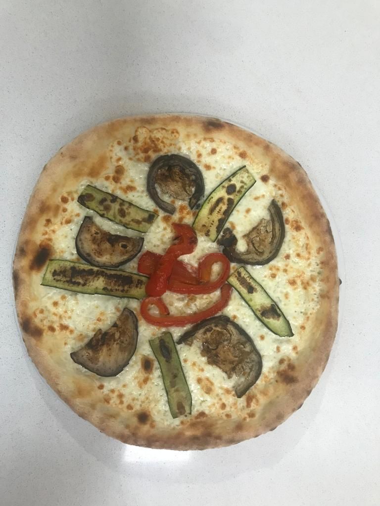 Una pizza vegetariana condita con zucchine grigliate, fette di melanzane e strisce di peperone rosso su una base bianca.