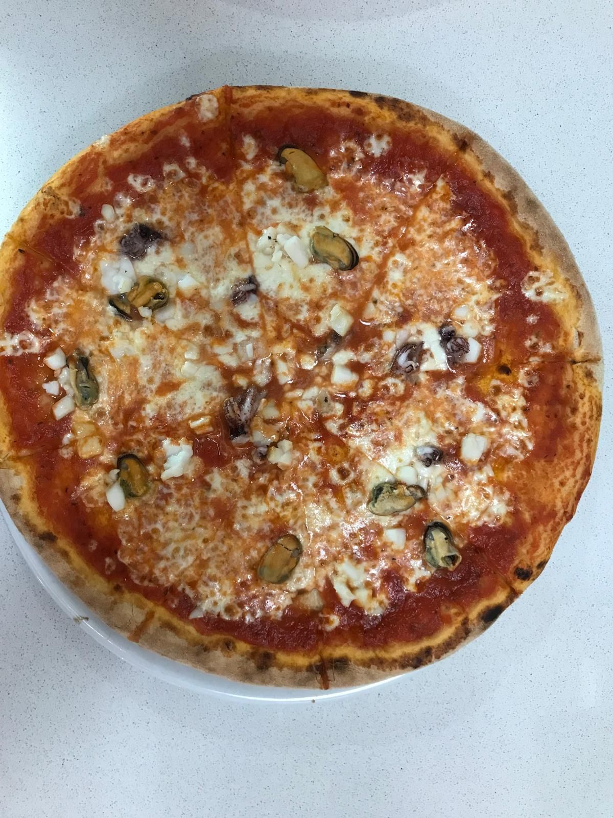 Una pizza rotonda a crosta sottile con salsa di pomodoro, formaggio bianco fuso e cozze nere sparse su un piatto bianco.