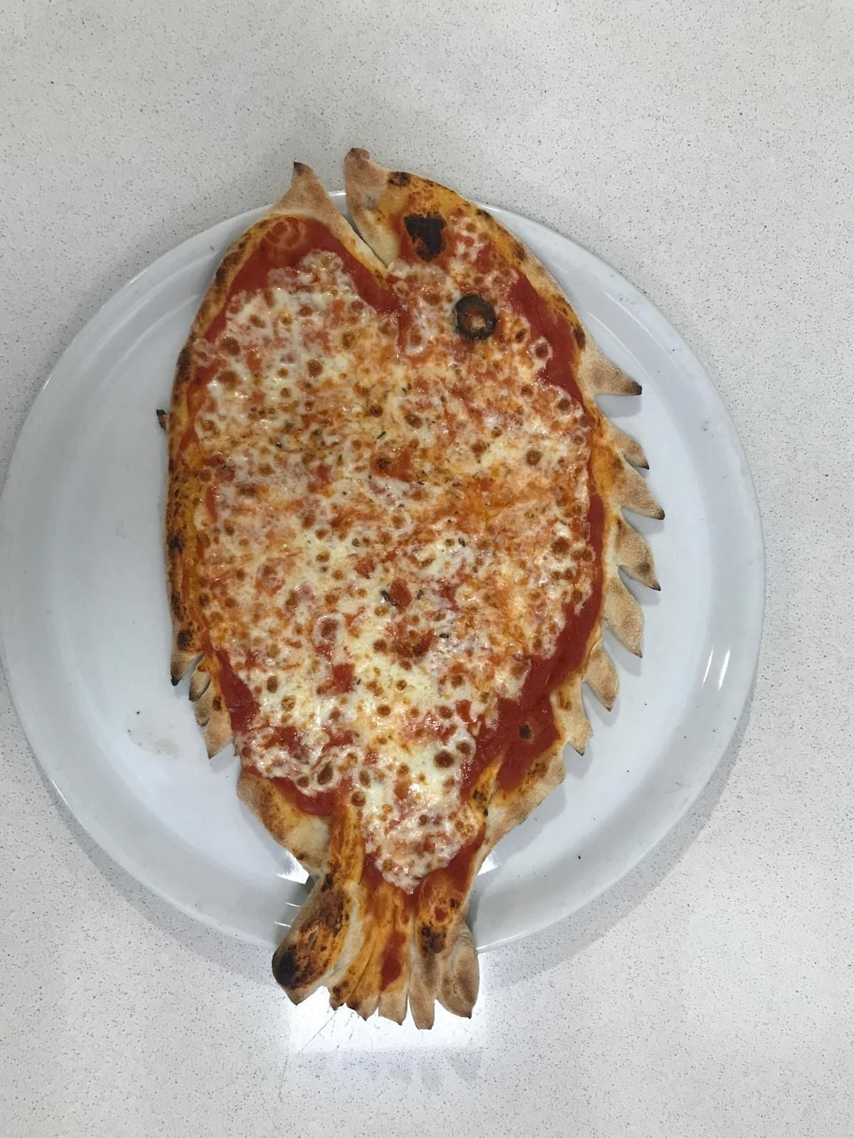 Una pizza a forma di pesce con salsa di pomodoro e formaggio fuso su un piatto bianco, appoggiata su un piano di lavoro maculato.