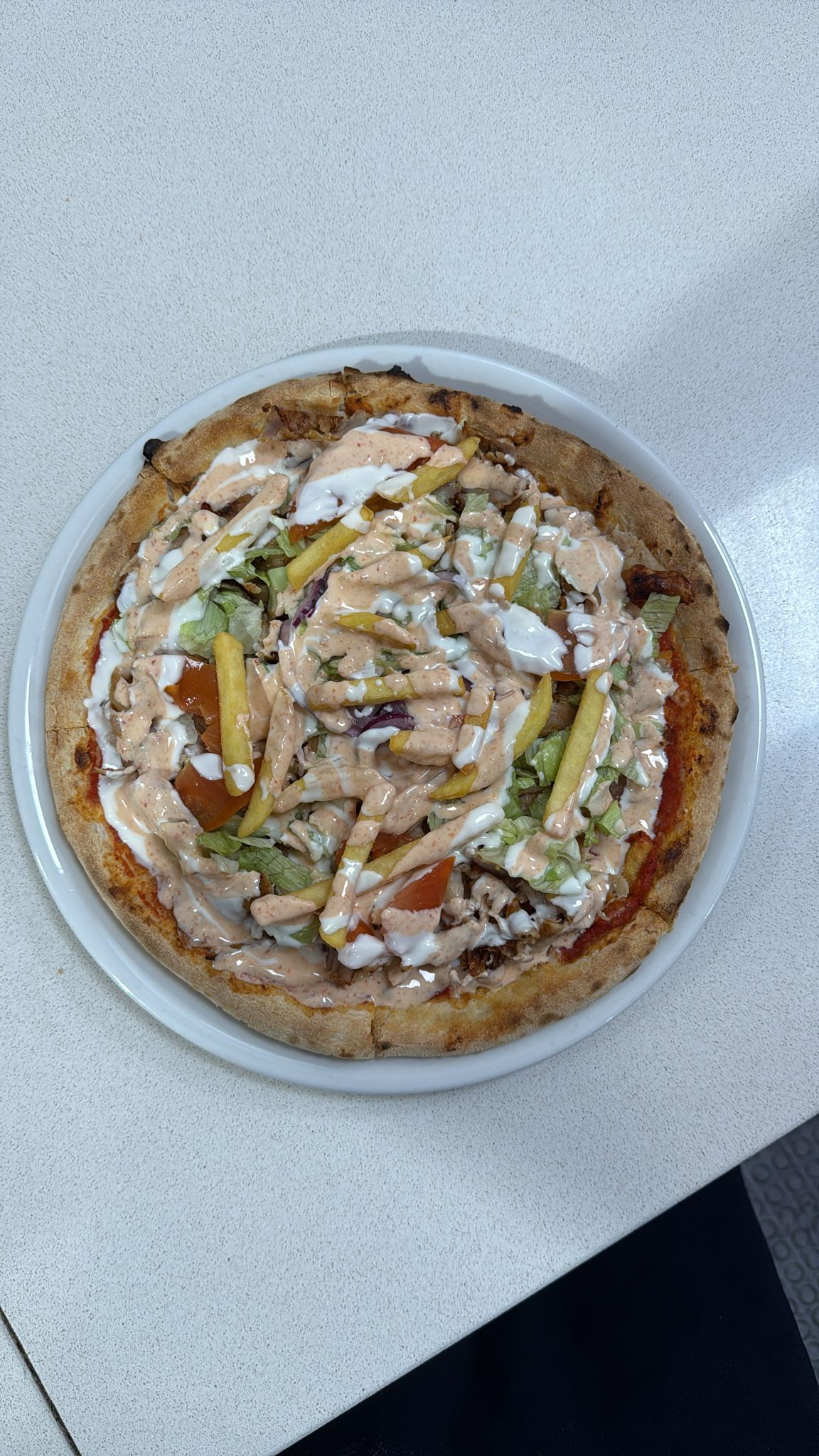 Una pizza su un piatto bianco, guarnita con lattuga tritata, patatine fritte e condita con salse bianca e rosa.