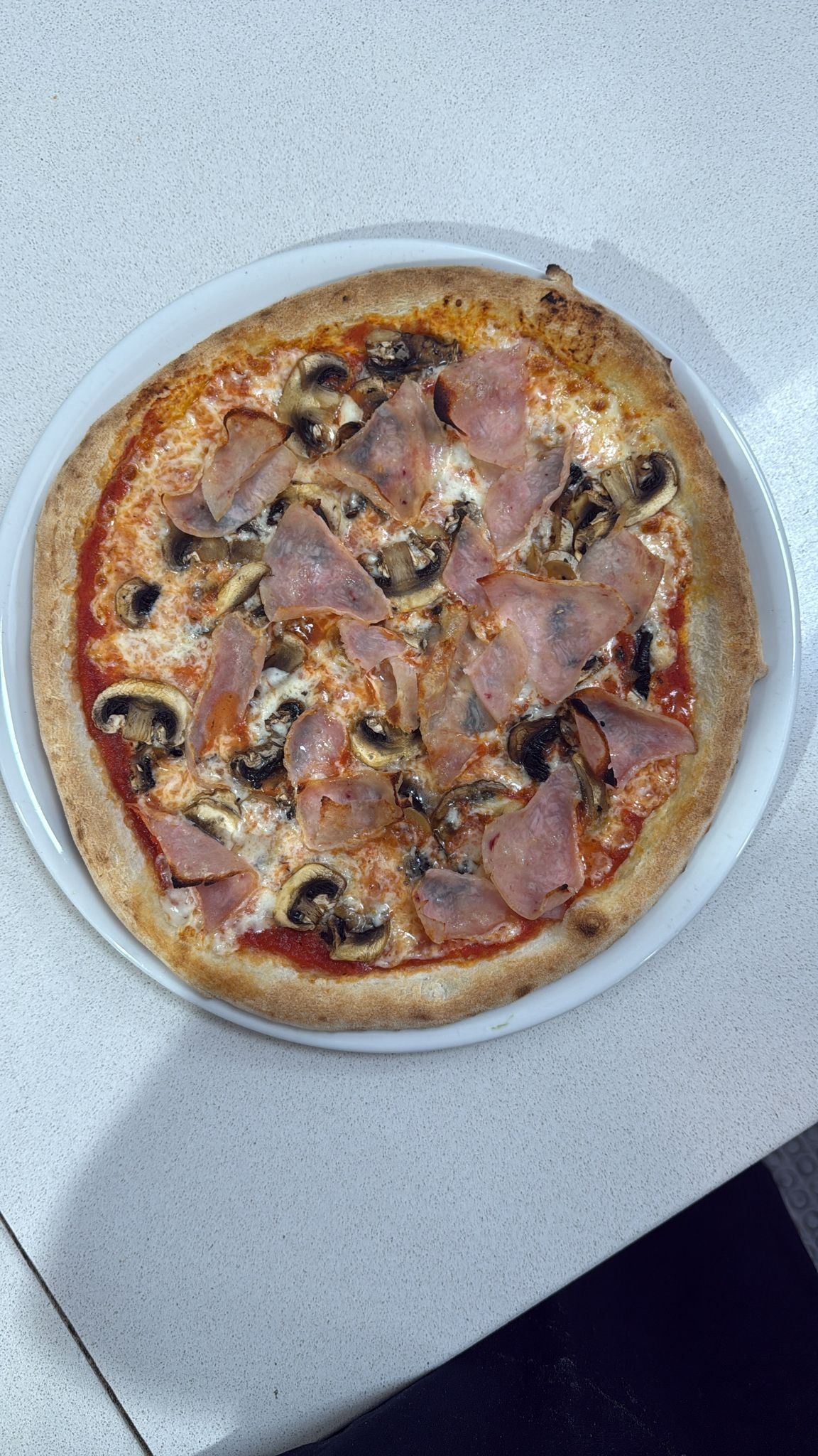 Una pizza rotonda con funghi e fette di prosciutto su una base di salsa di pomodoro, posizionata al centro di un piatto bianco.