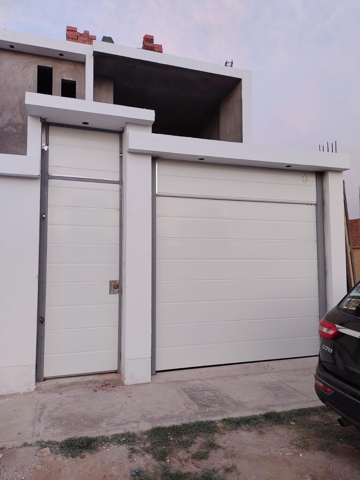Puerta de garaje blanca y portón peatonal frente a una casa a medio construir. Un coche oscuro está aparcado a la derecha.