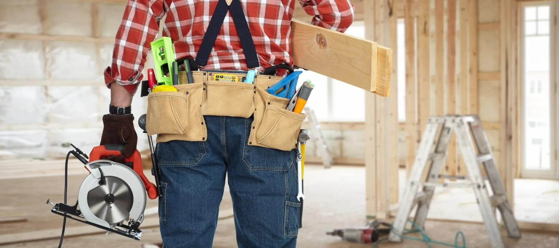 A man is holding a circular saw and a piece of wood in a room under construction.