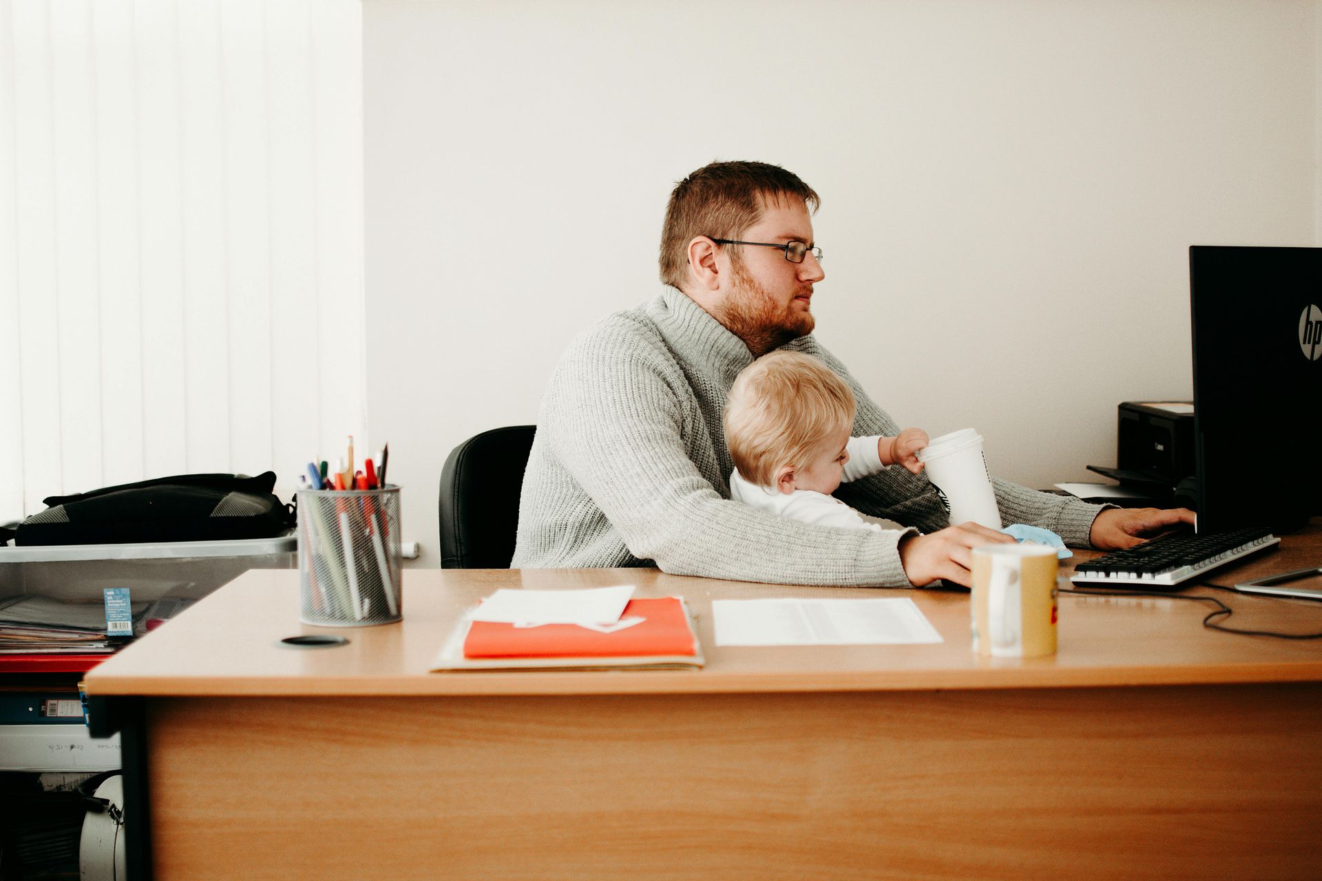 Man with child on lap submitting a quote of graded sports cards at a computer in an office, beige sweater, desk.
