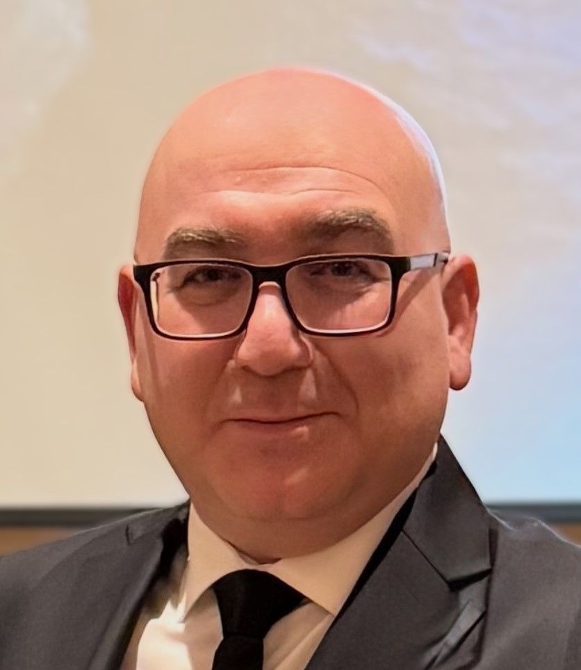 A bald person with glasses wearing a black suit and tie, smiling slightly against a neutral background.